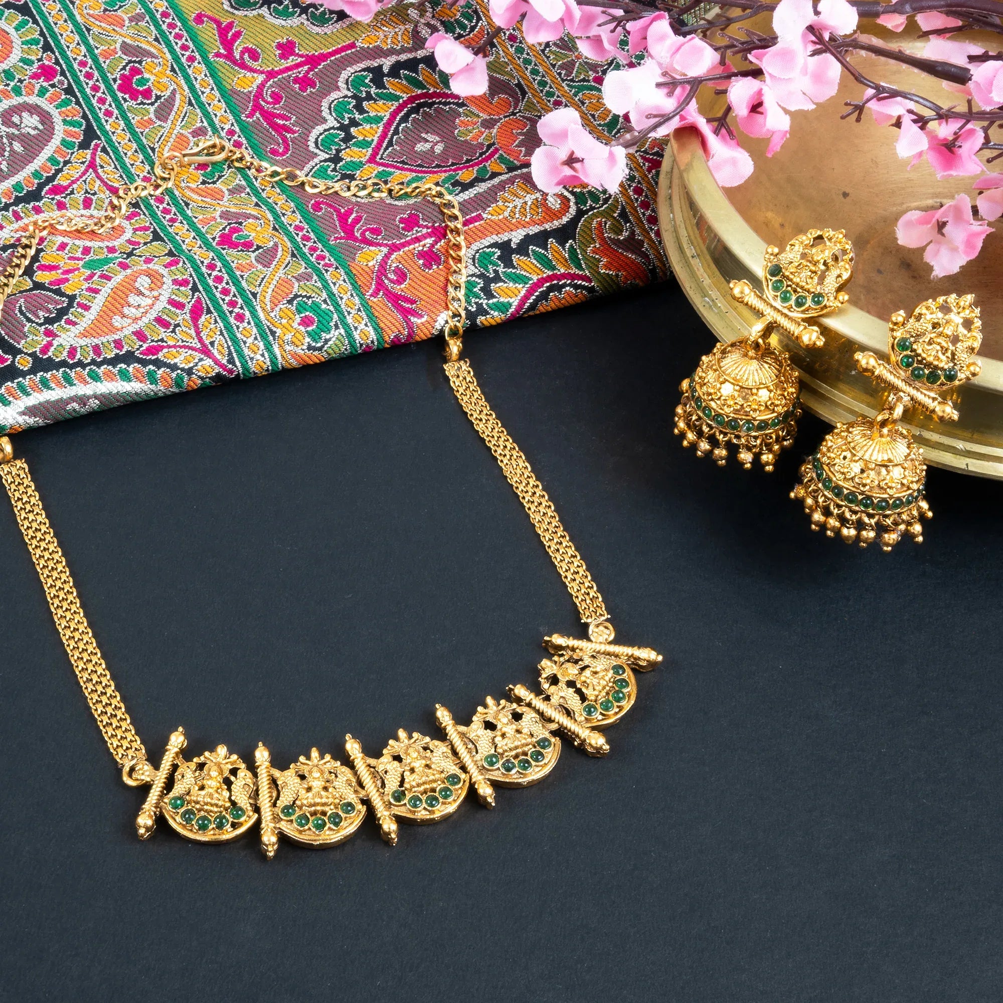 Gold necklace and earrings on a dark surface with a colorful fabric and pink flowers in the background.