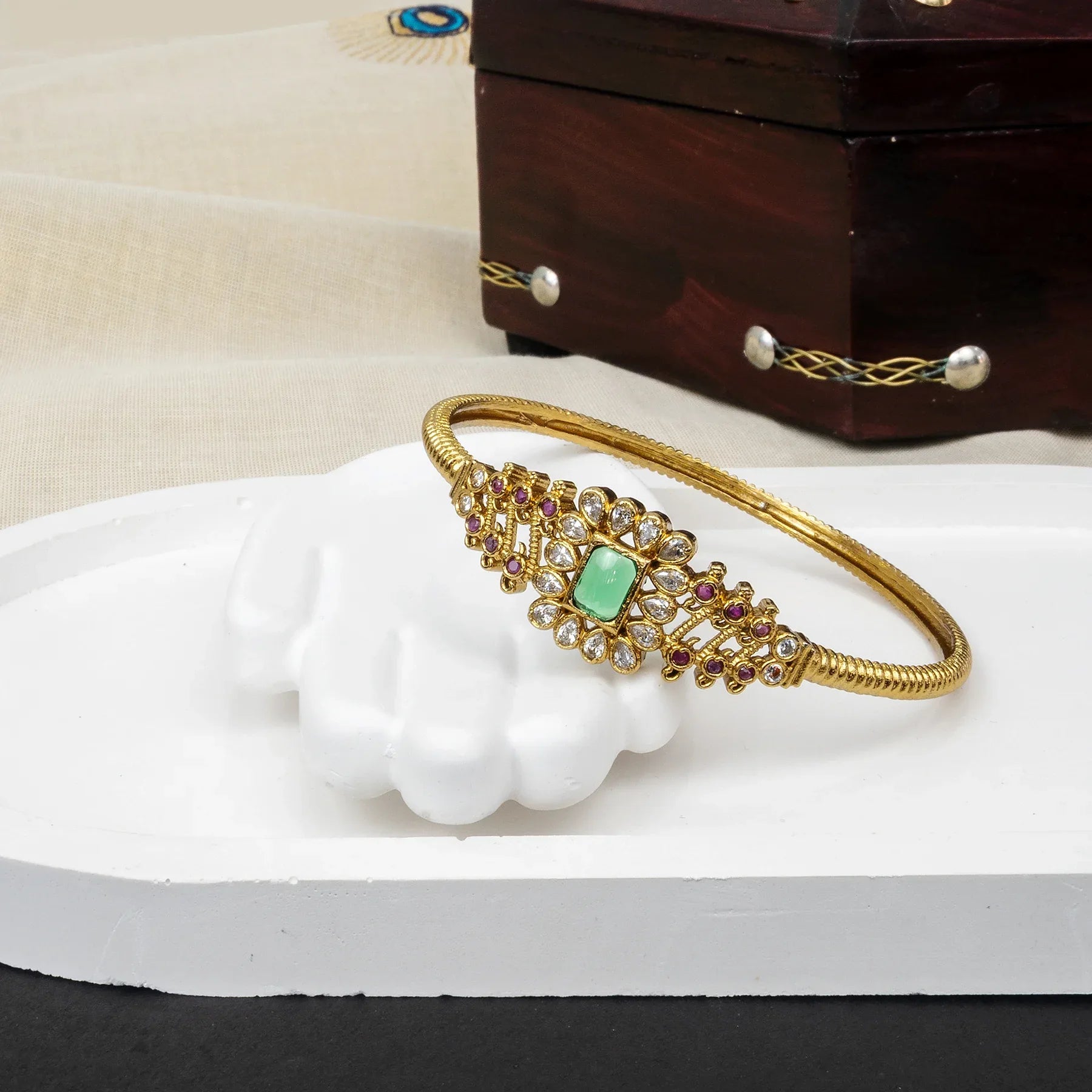 Gold bracelet with green gemstone on a white surface with a wooden box in the background