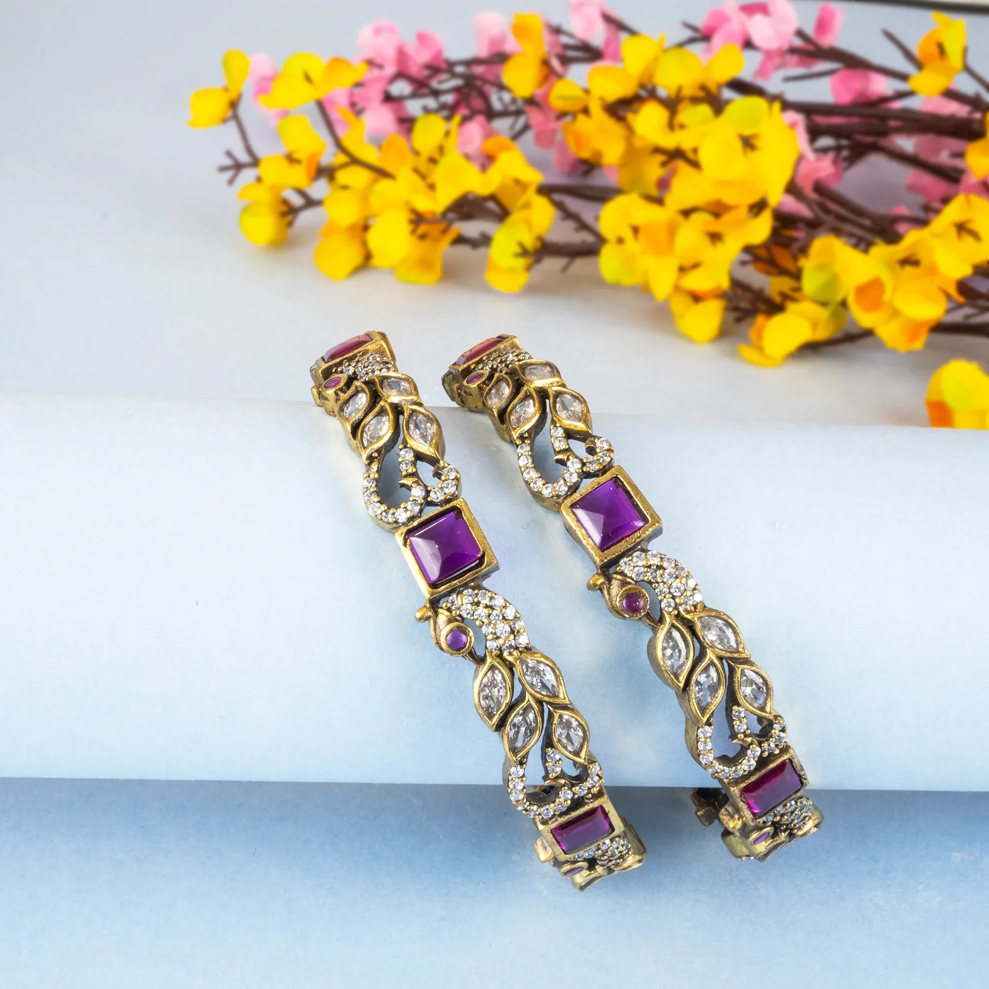 Gold bangles with purple stones on a white surface with yellow flowers in the background