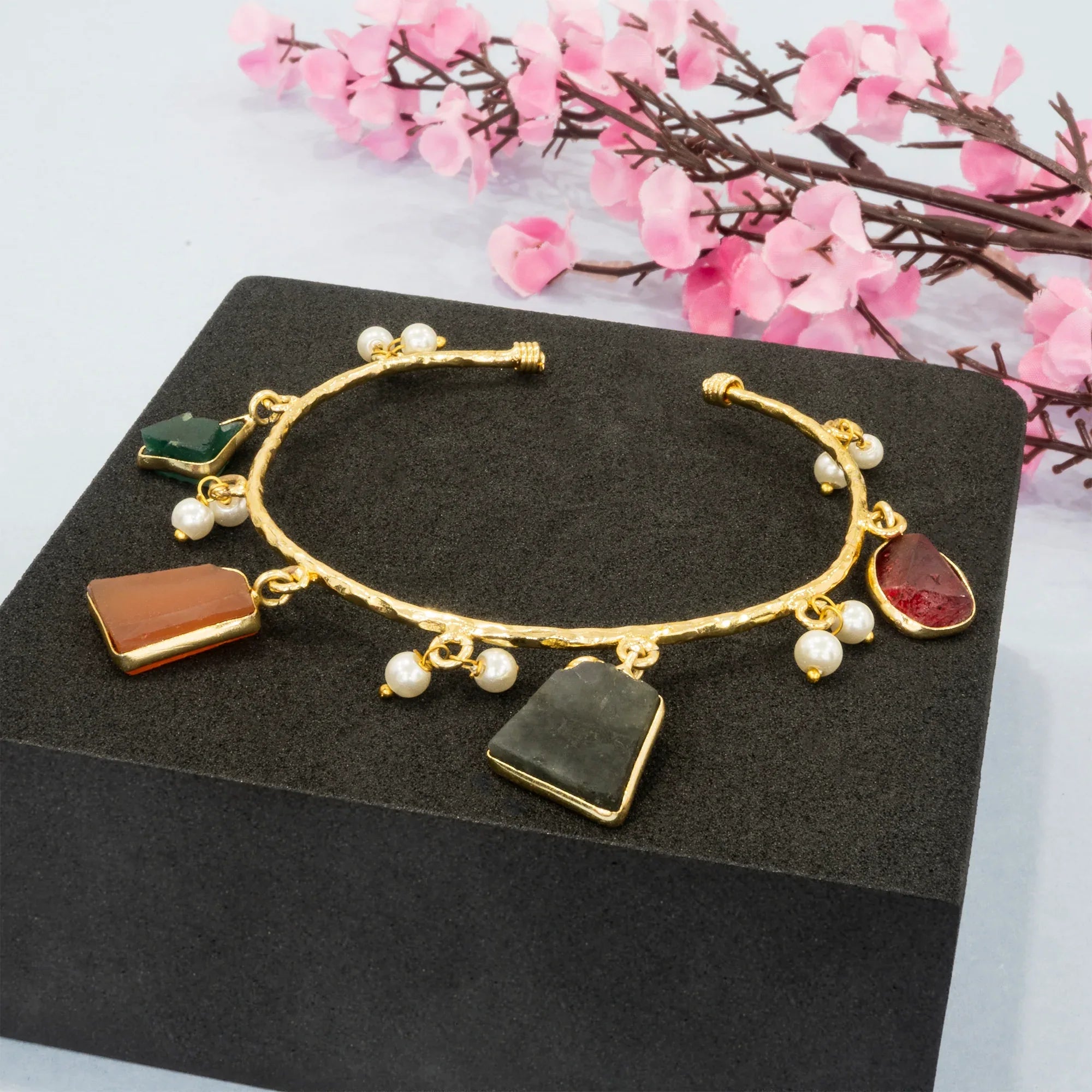 Gold bracelet with various charms on a black stand with pink flowers in the background