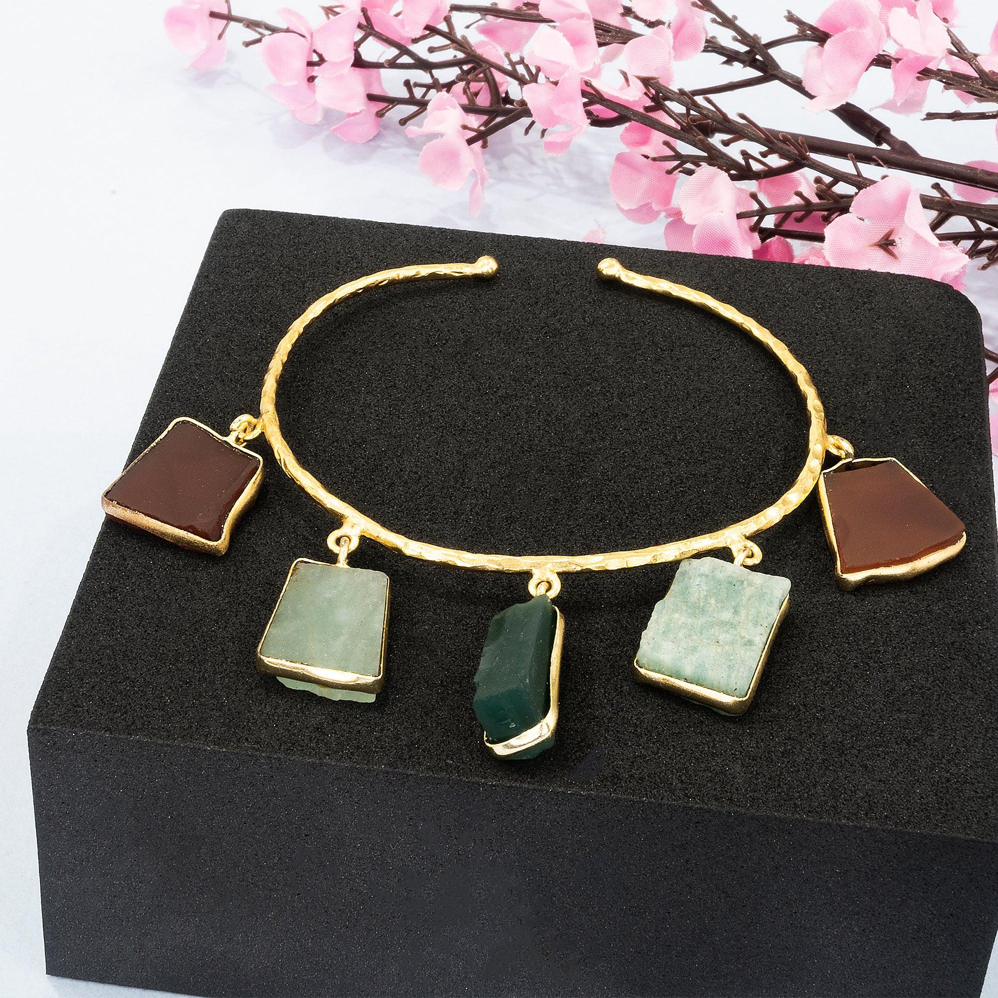 Gold bracelet with green stone pendants on a black stand with pink flowers in the background