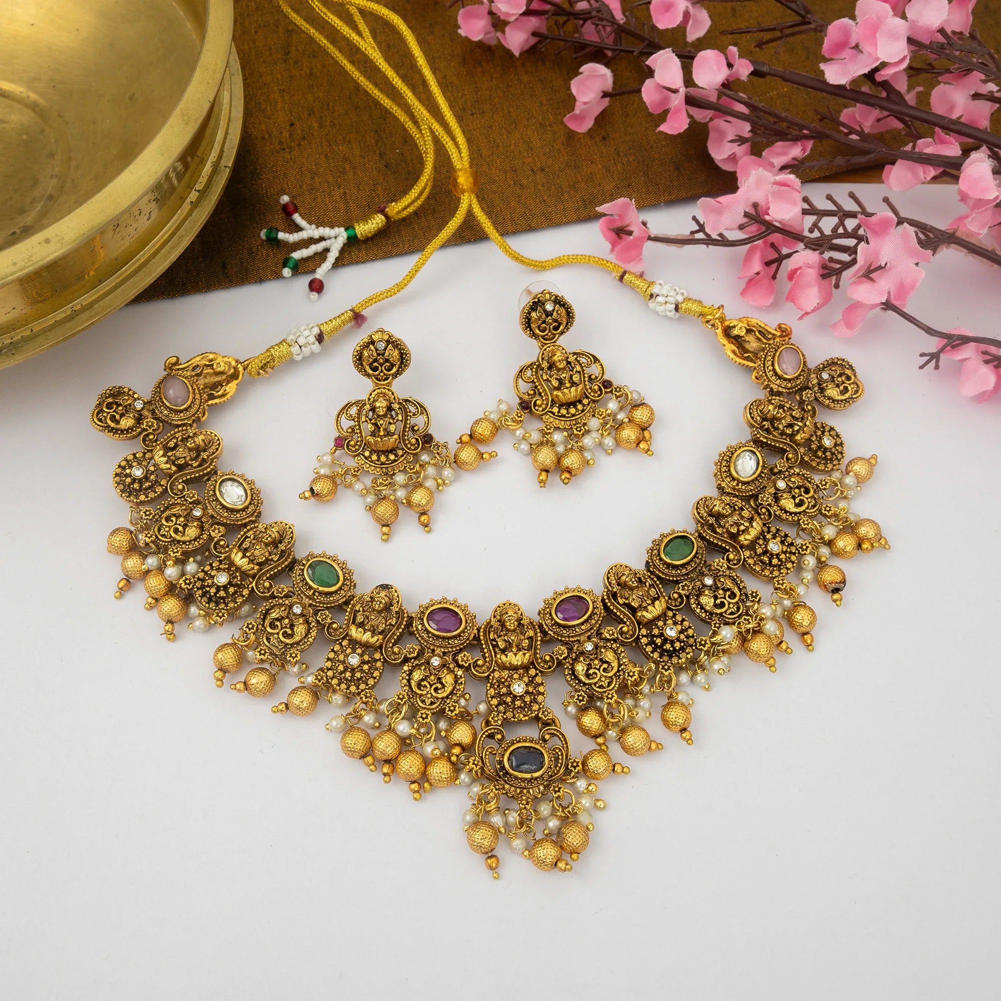 Gold necklace and earrings with gemstones on a white surface with pink flowers in the background