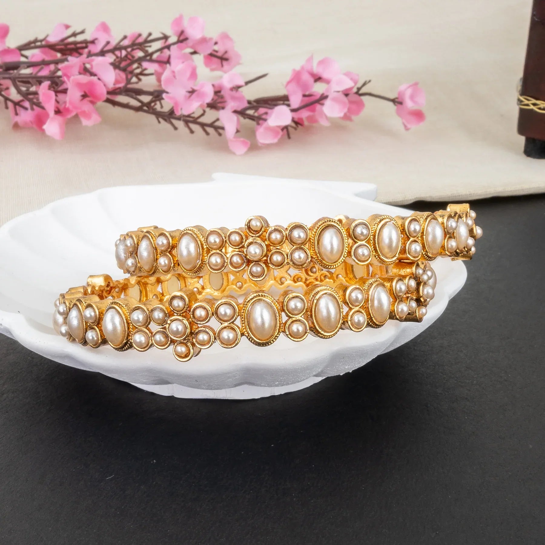 Gold and pearl bracelet on a white dish with pink flowers in the background