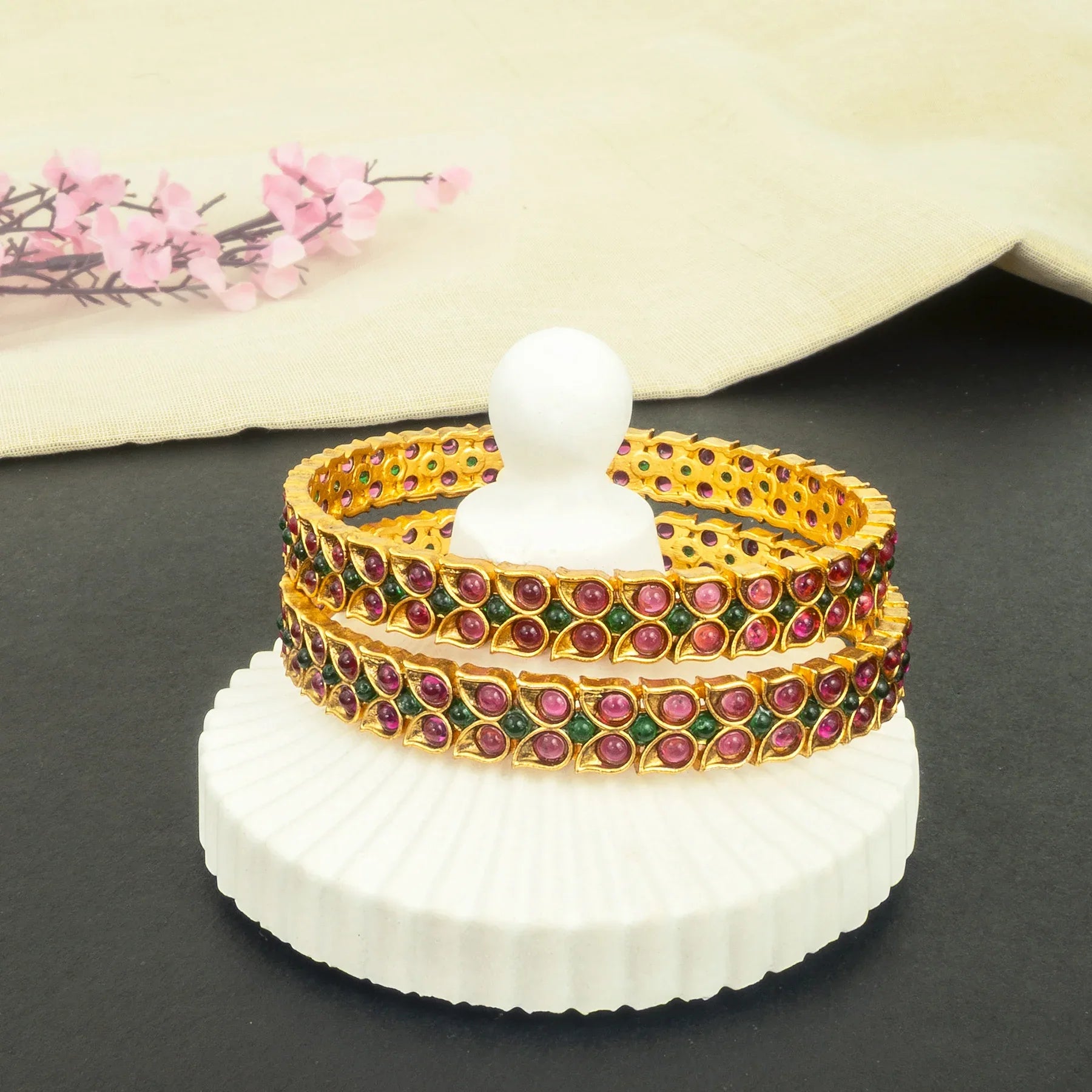 Set of gold bangles with red and green stones on a white stand against a neutral background