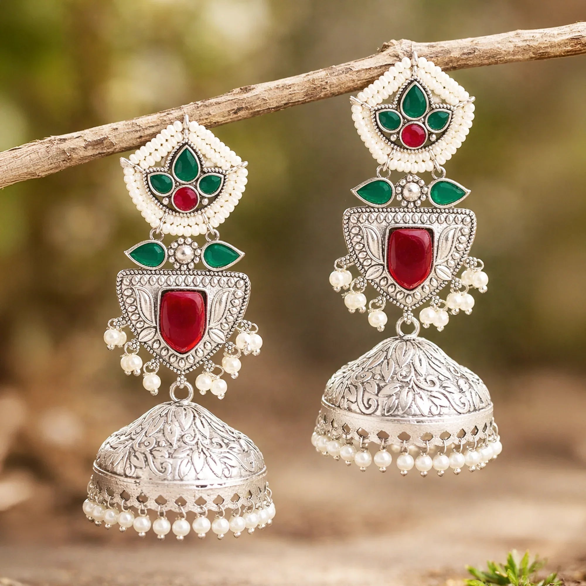 Ornate silver earrings with red and green stones on a branch