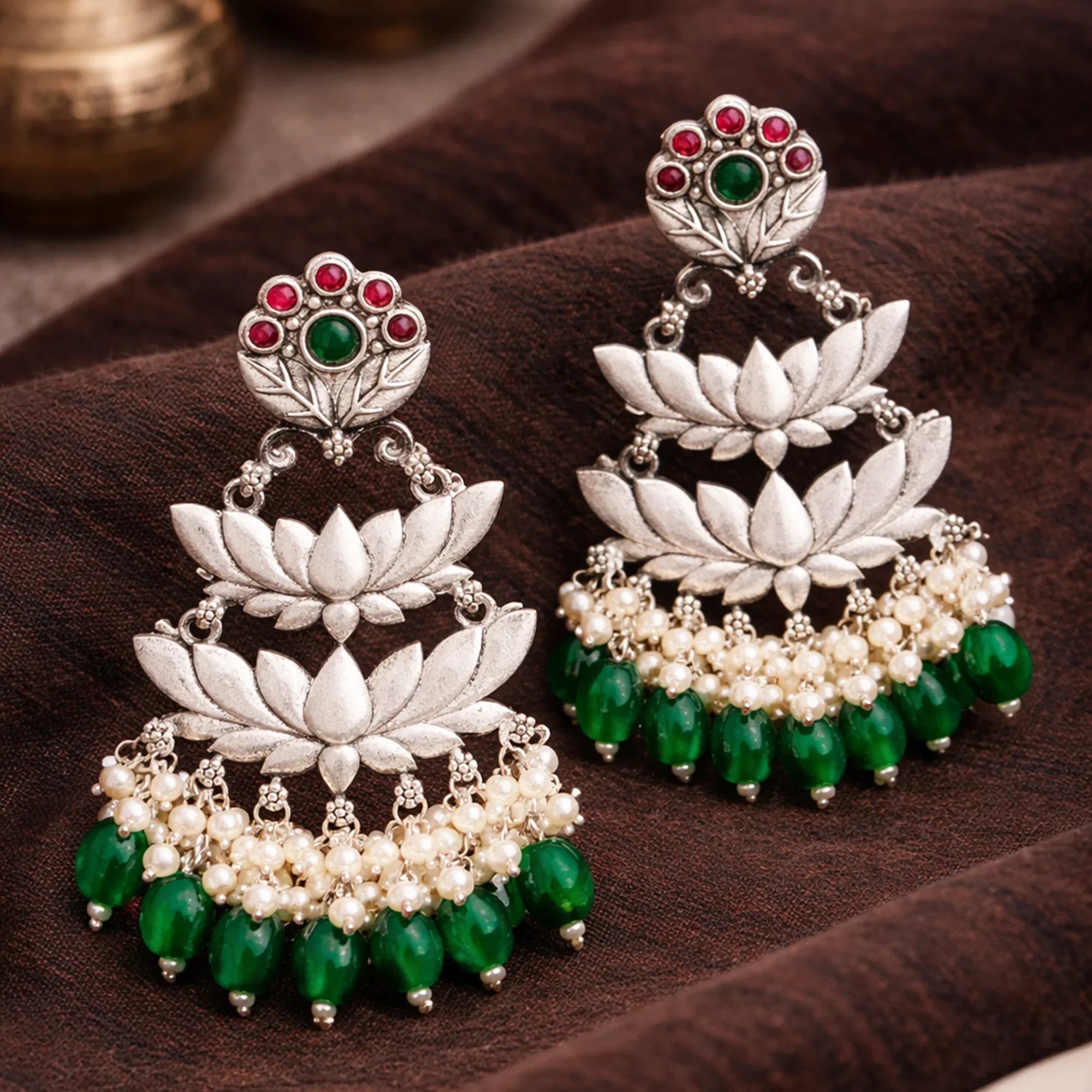 Ornate silver earrings with green and red gemstones on a brown fabric background