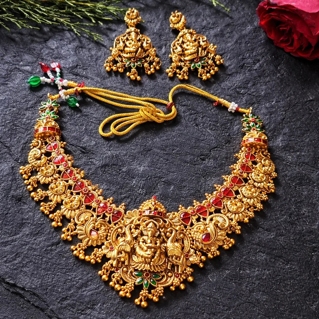 Gold necklace and earrings on a dark surface with green leaves and a red rose.