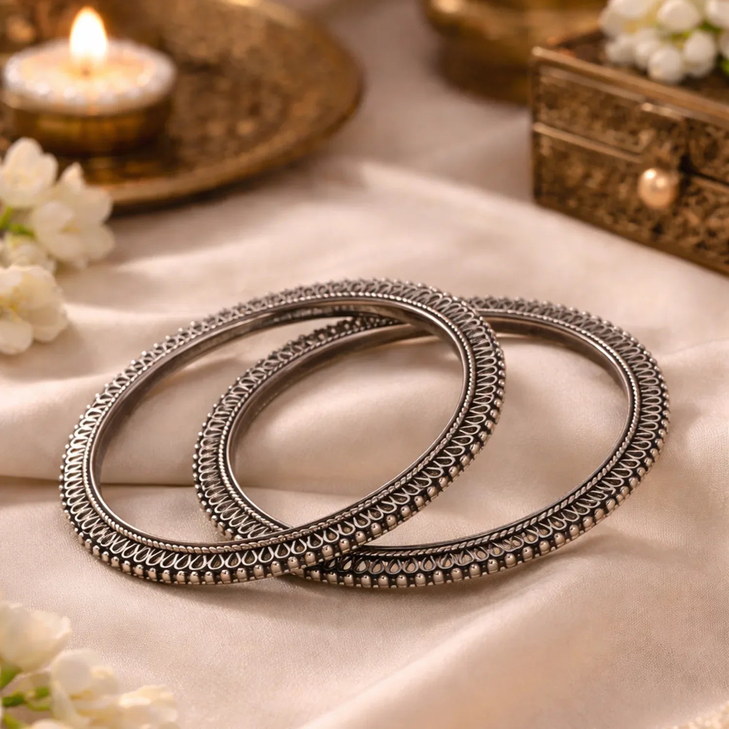 Pair of silver bangles on a soft fabric background with candles and flowers.