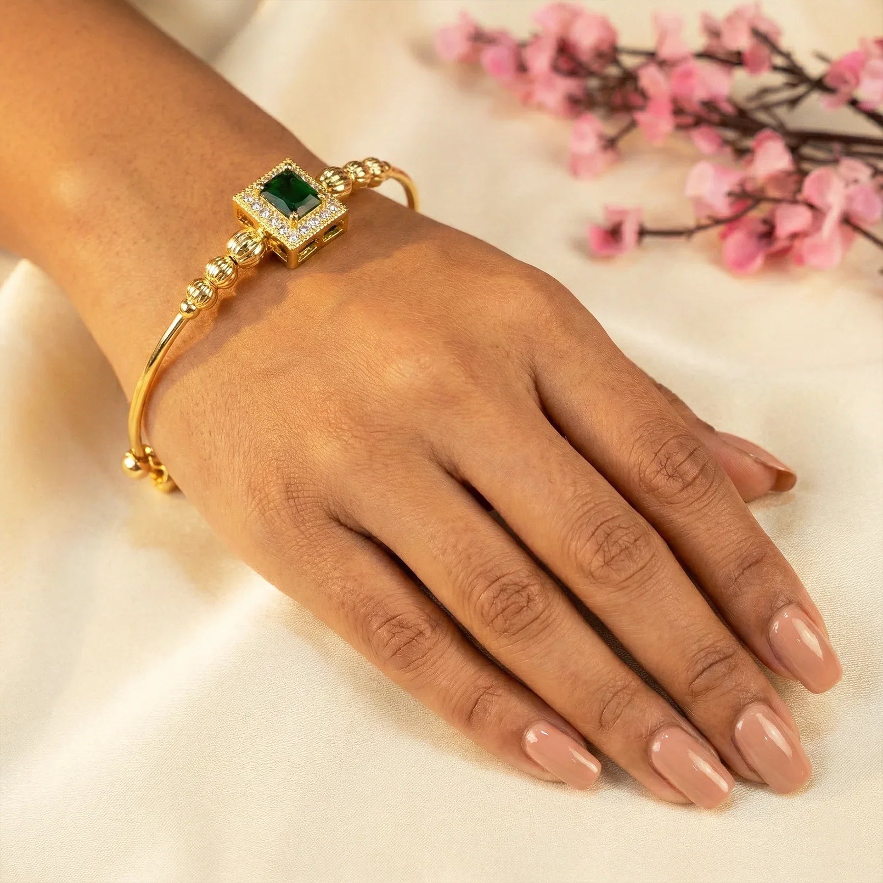 Gold bracelet with a green gemstone on a hand against a light background with pink flowers.