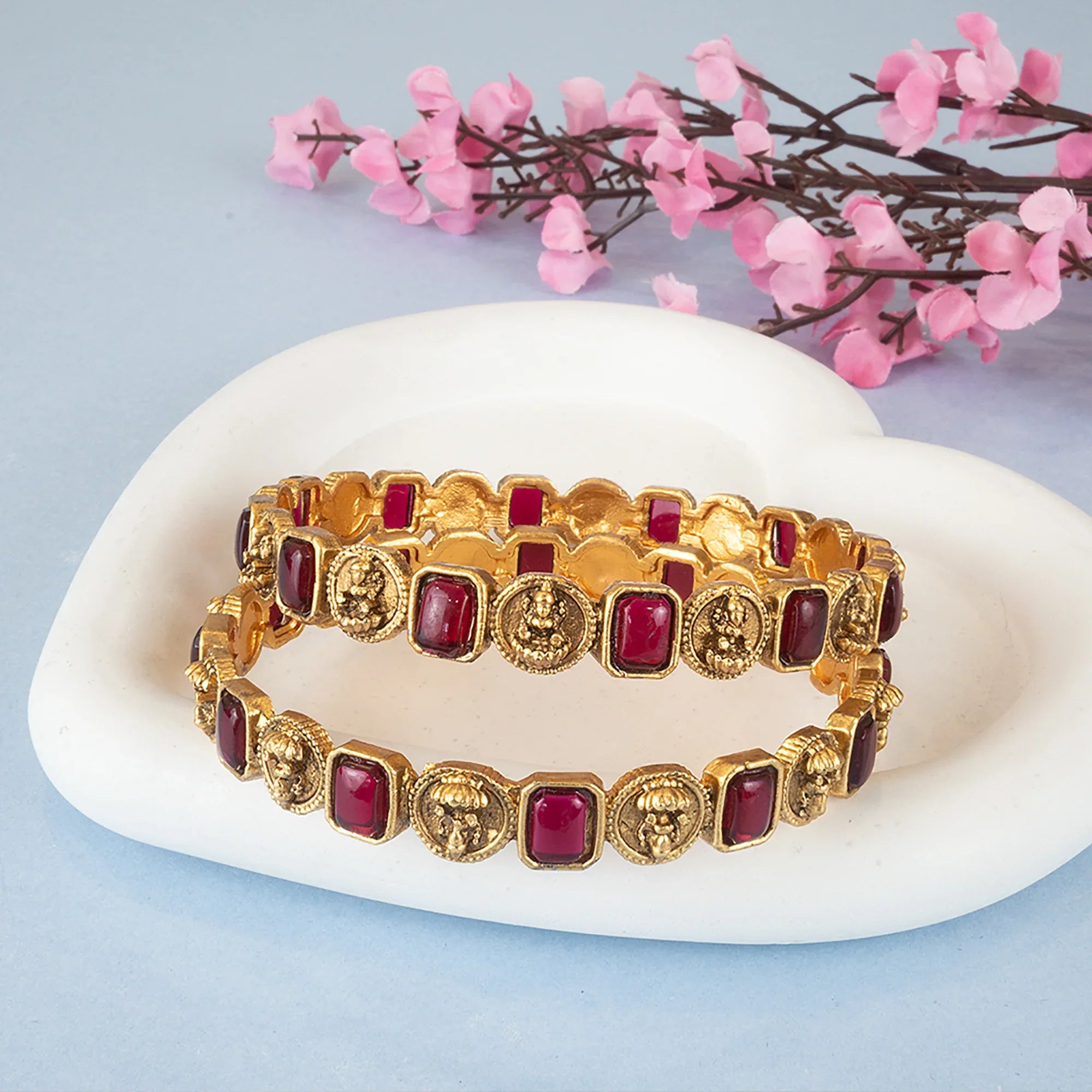 Gold bracelets with red stones on a white dish with pink flowers in the background