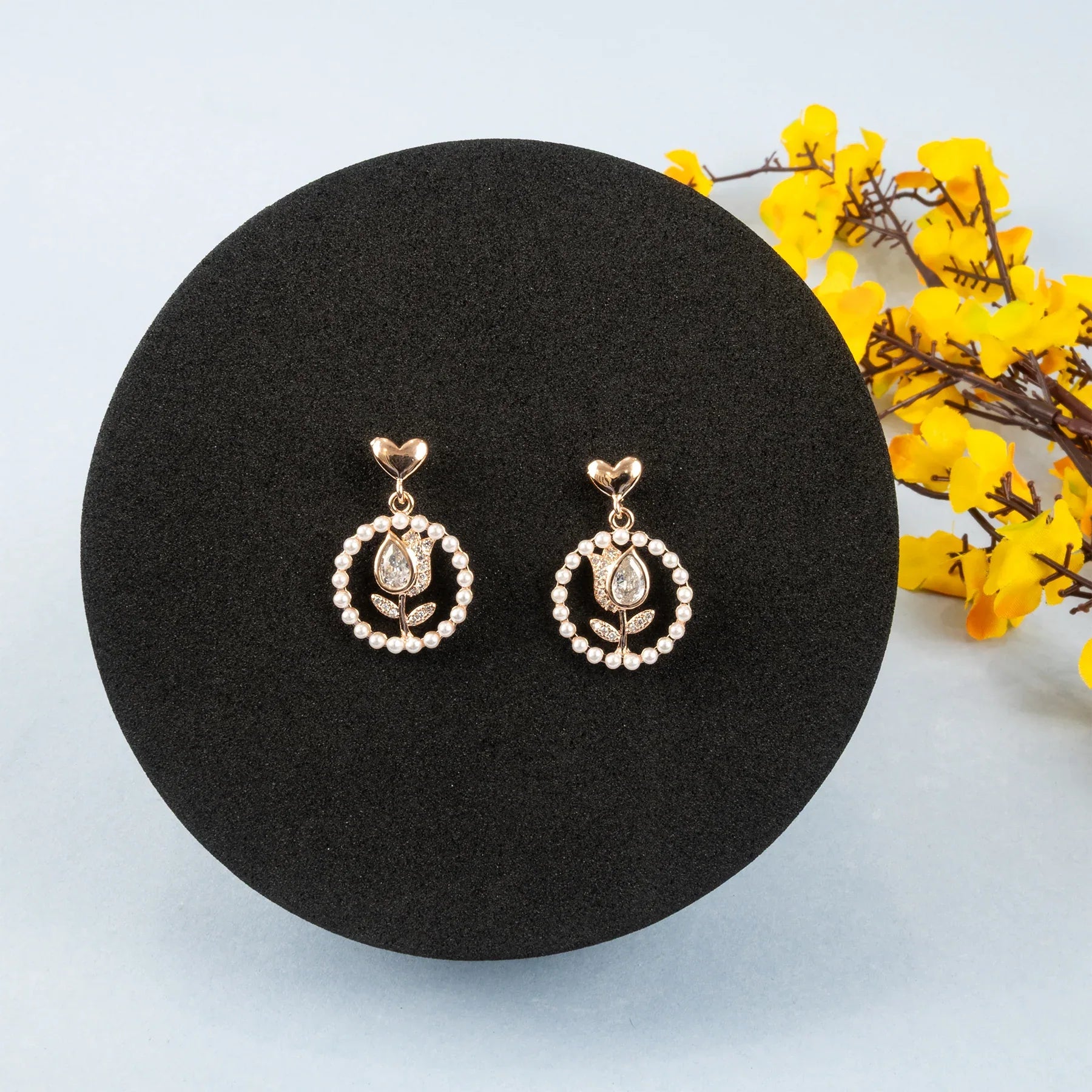Pair of gold and pearl earrings on a black round stand with yellow flowers in the background.