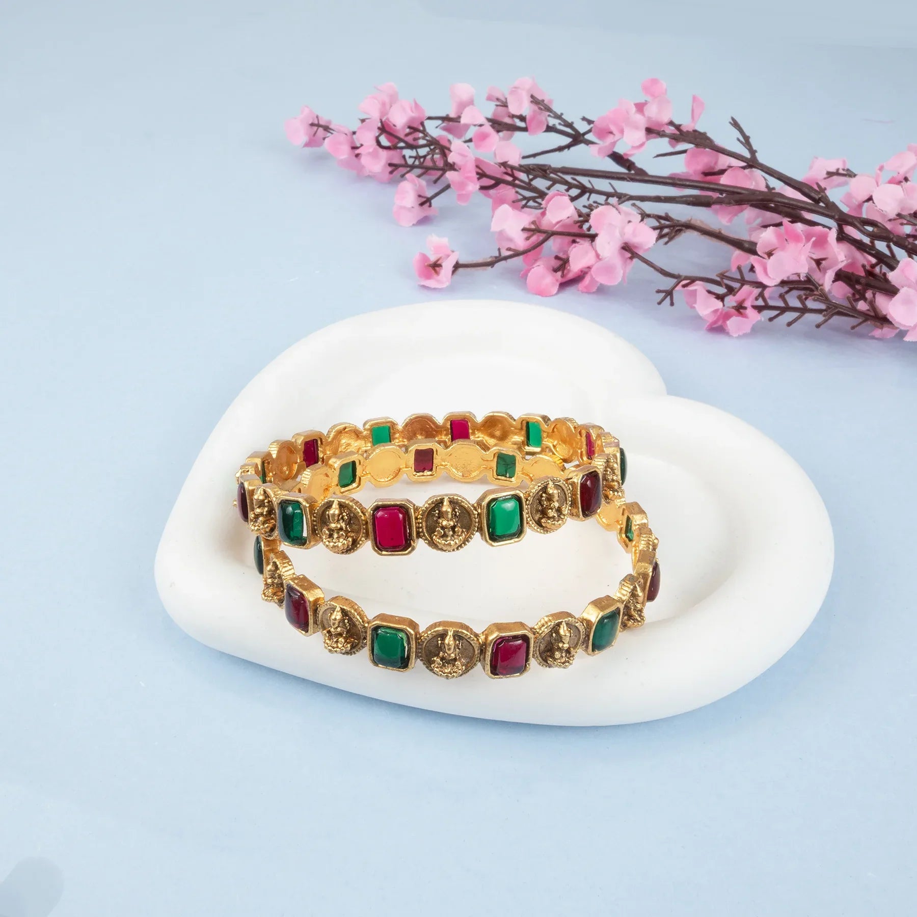 Gold bracelets with colorful stones on a white stone surface with pink flowers in the background