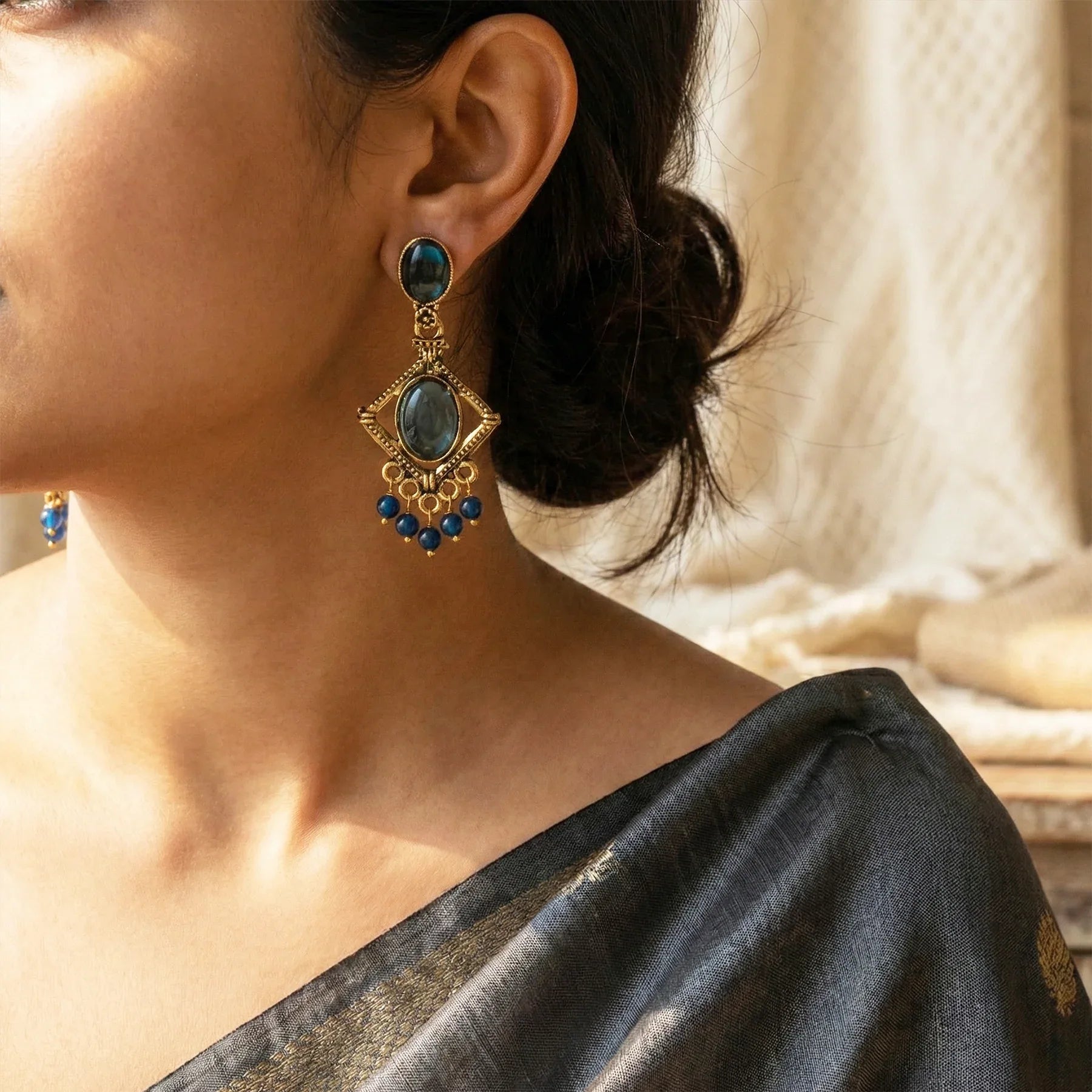 Close-up of a person wearing gold earrings with blue stones, set against a neutral background.