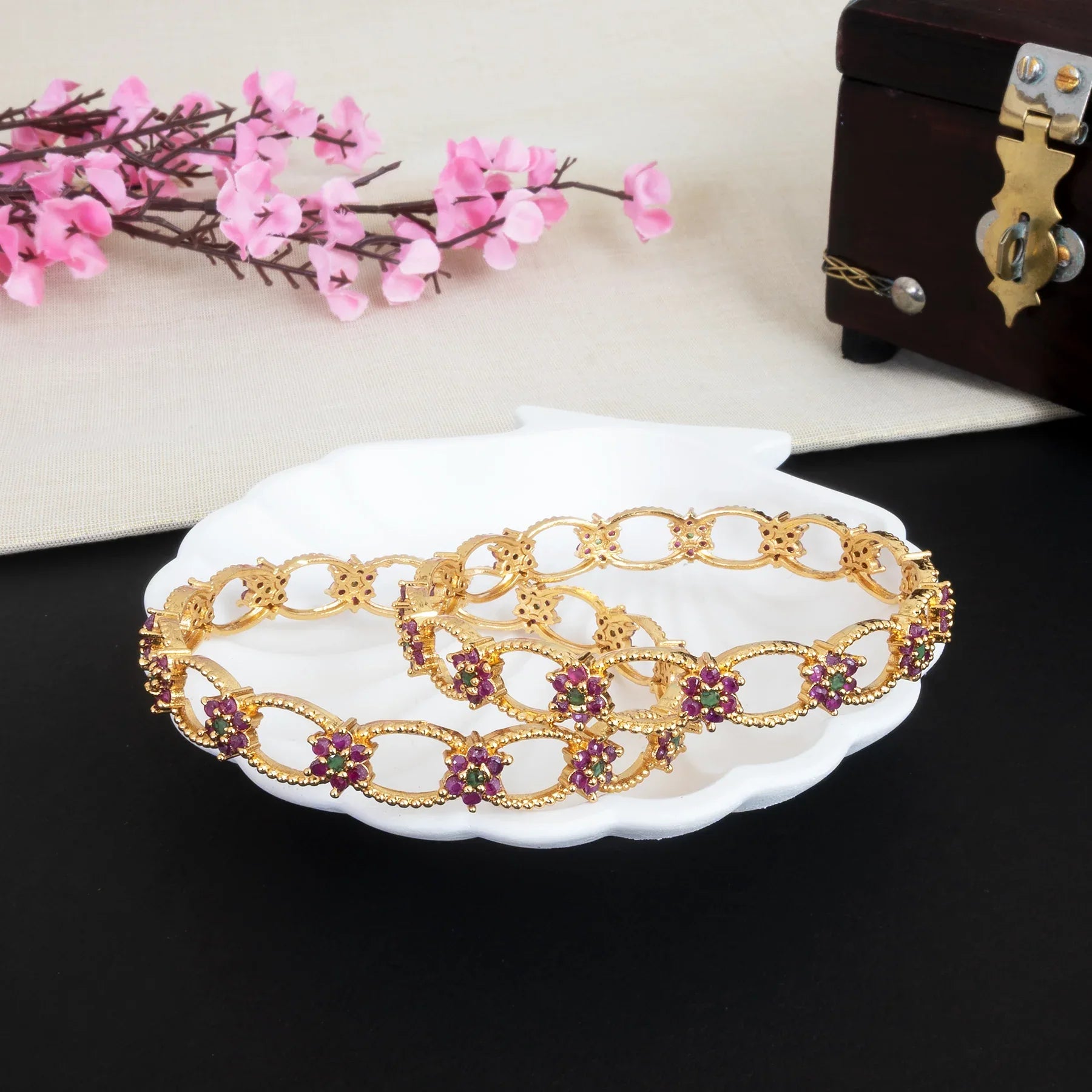 Decorative gold bracelet with gemstones on a white dish, with pink flowers and a wooden box in the background.