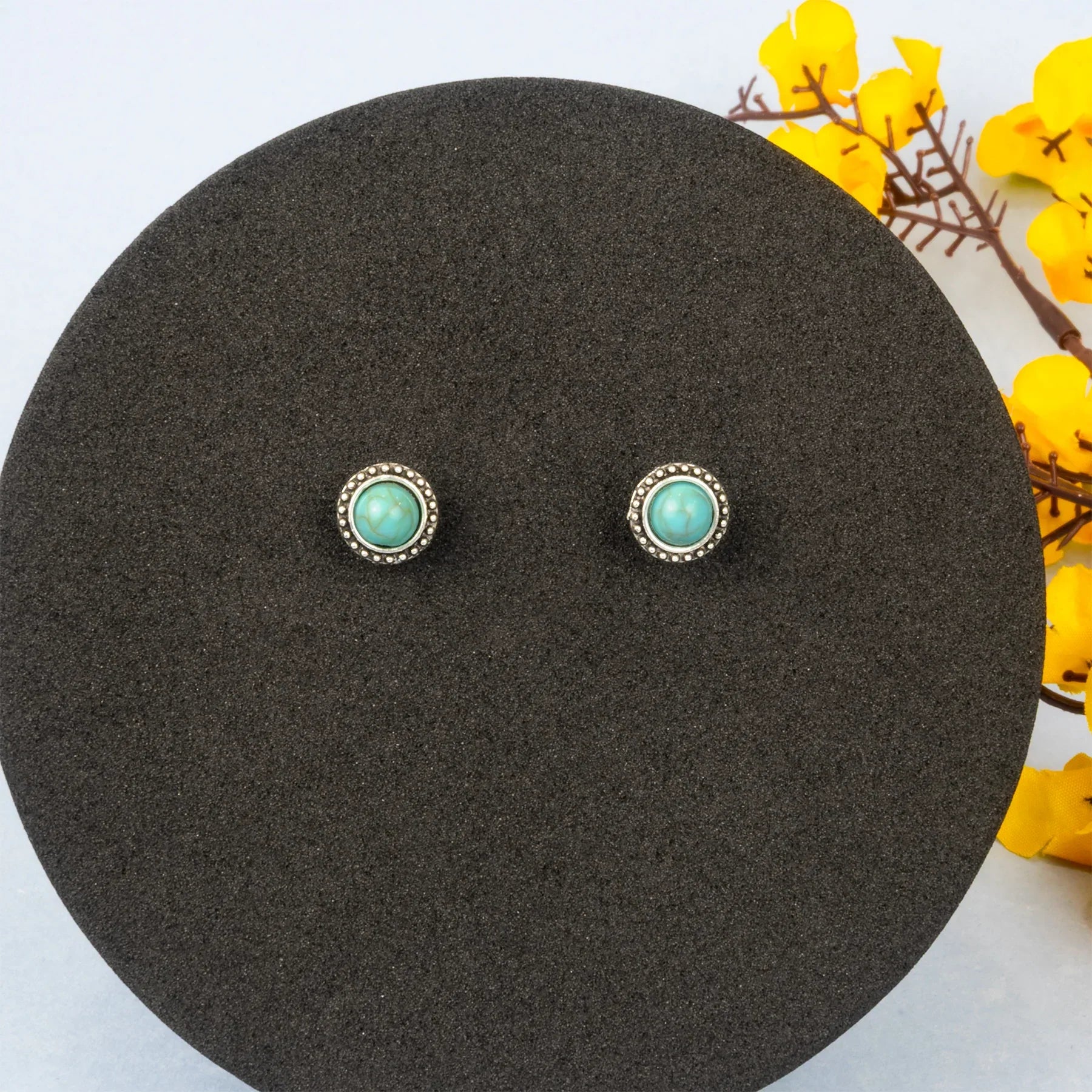Pair of turquoise stud earrings on a black round stand with yellow flowers in the background.