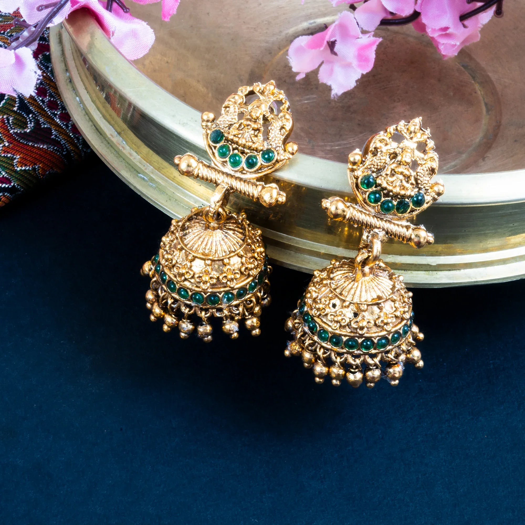 Gold earrings with green gemstones on a reflective surface with pink flowers.