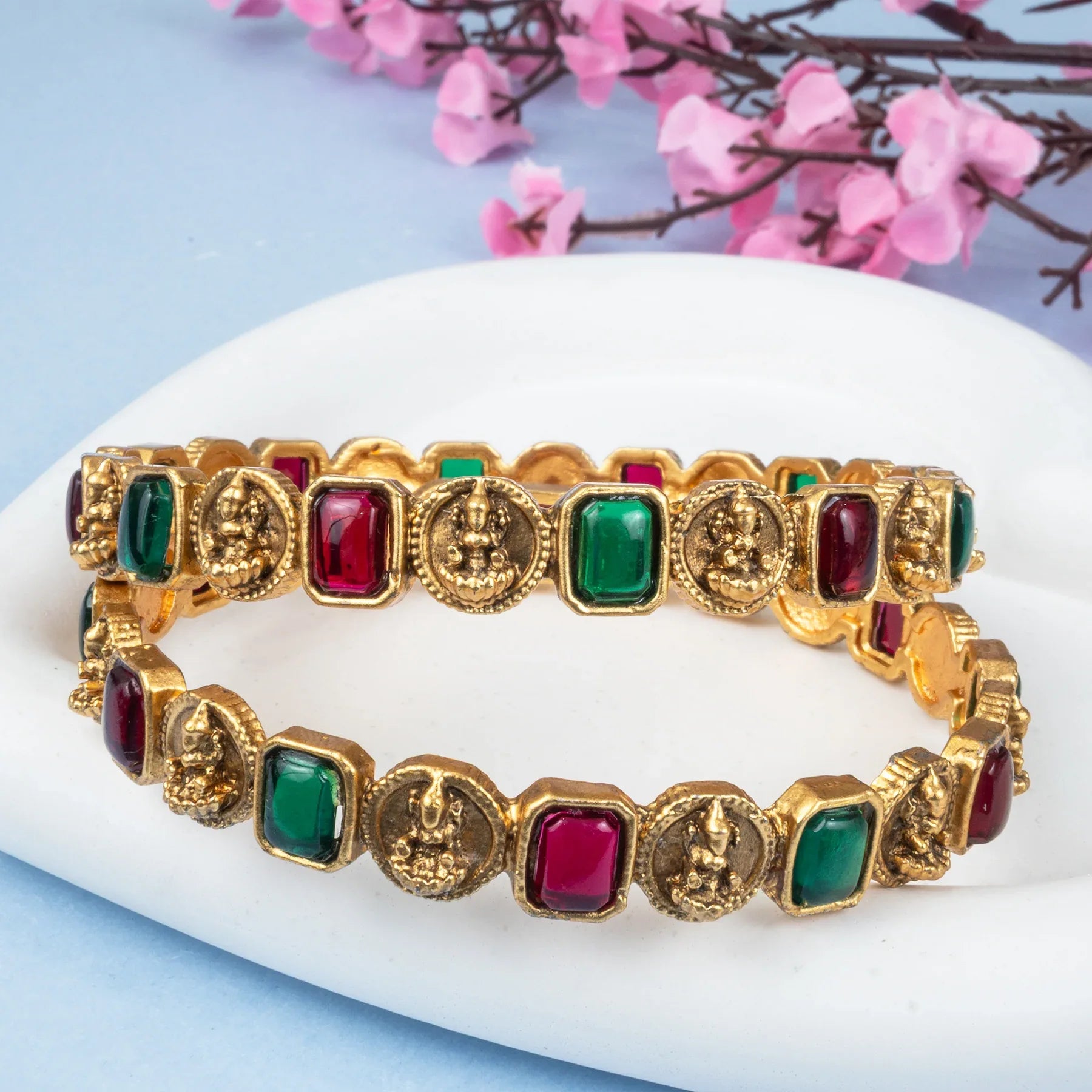 Gold bracelets with red and green stones on a white surface with pink flowers in the background