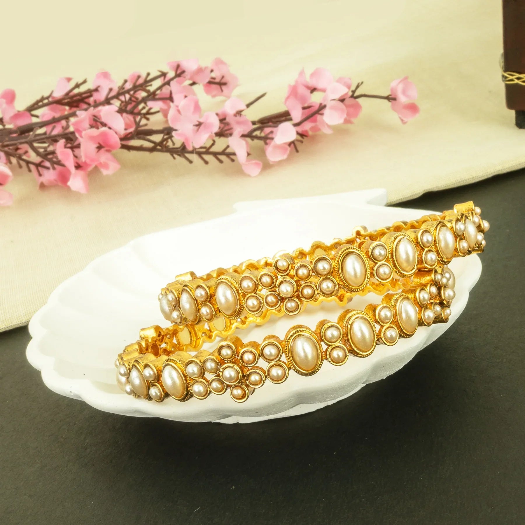 Gold and pearl bangles on a white dish with pink flowers in the background