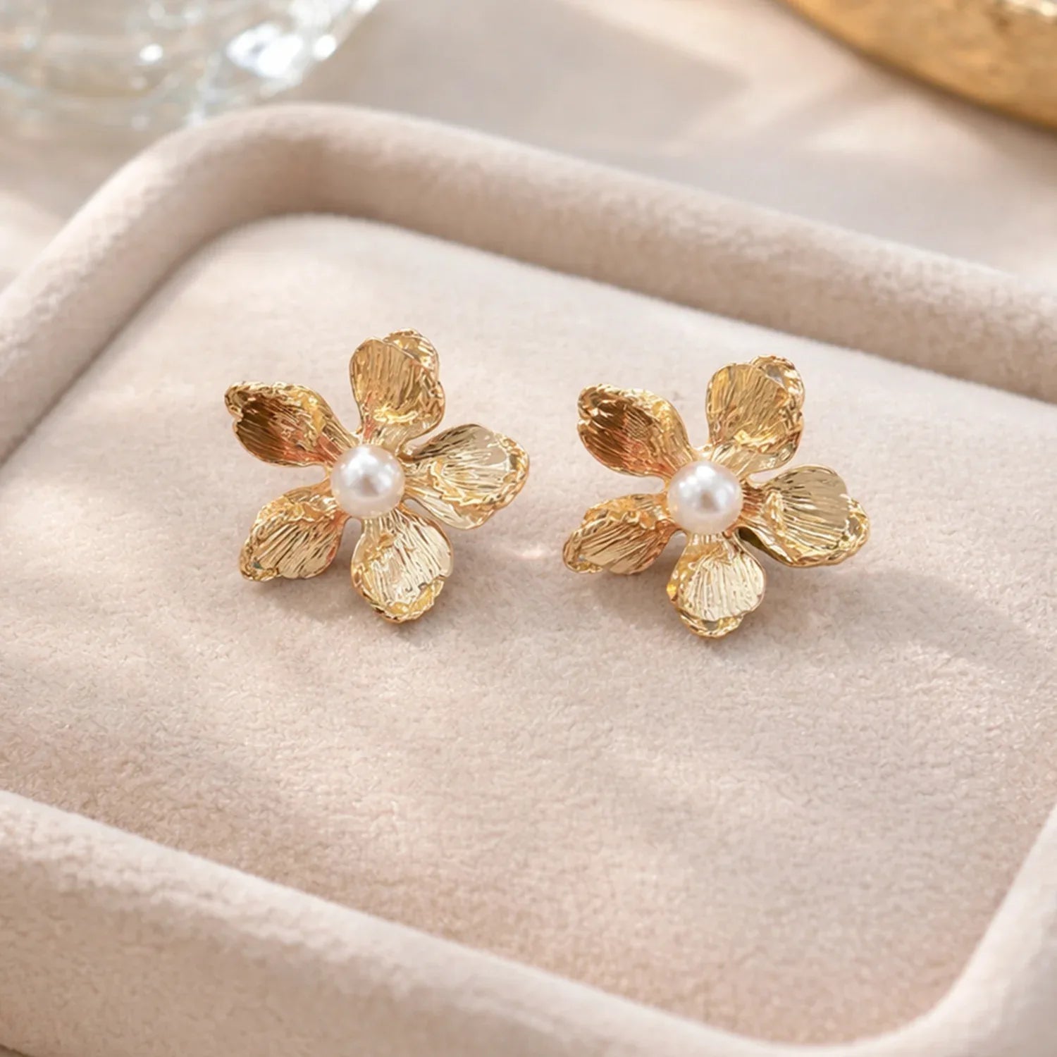 Gold flower-shaped earrings with pearl centers on a beige fabric background