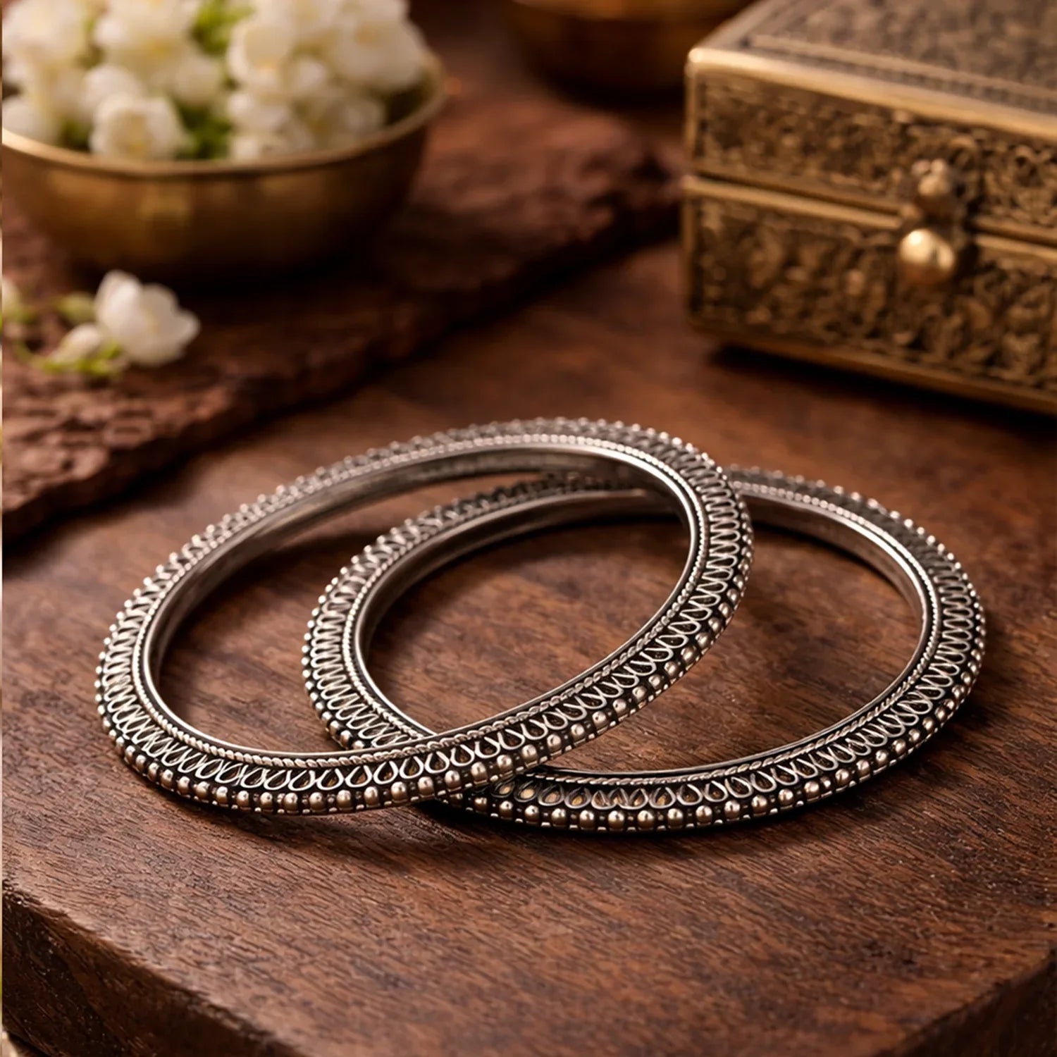Two silver bangles on a wooden surface with a decorative box and flowers in the background.