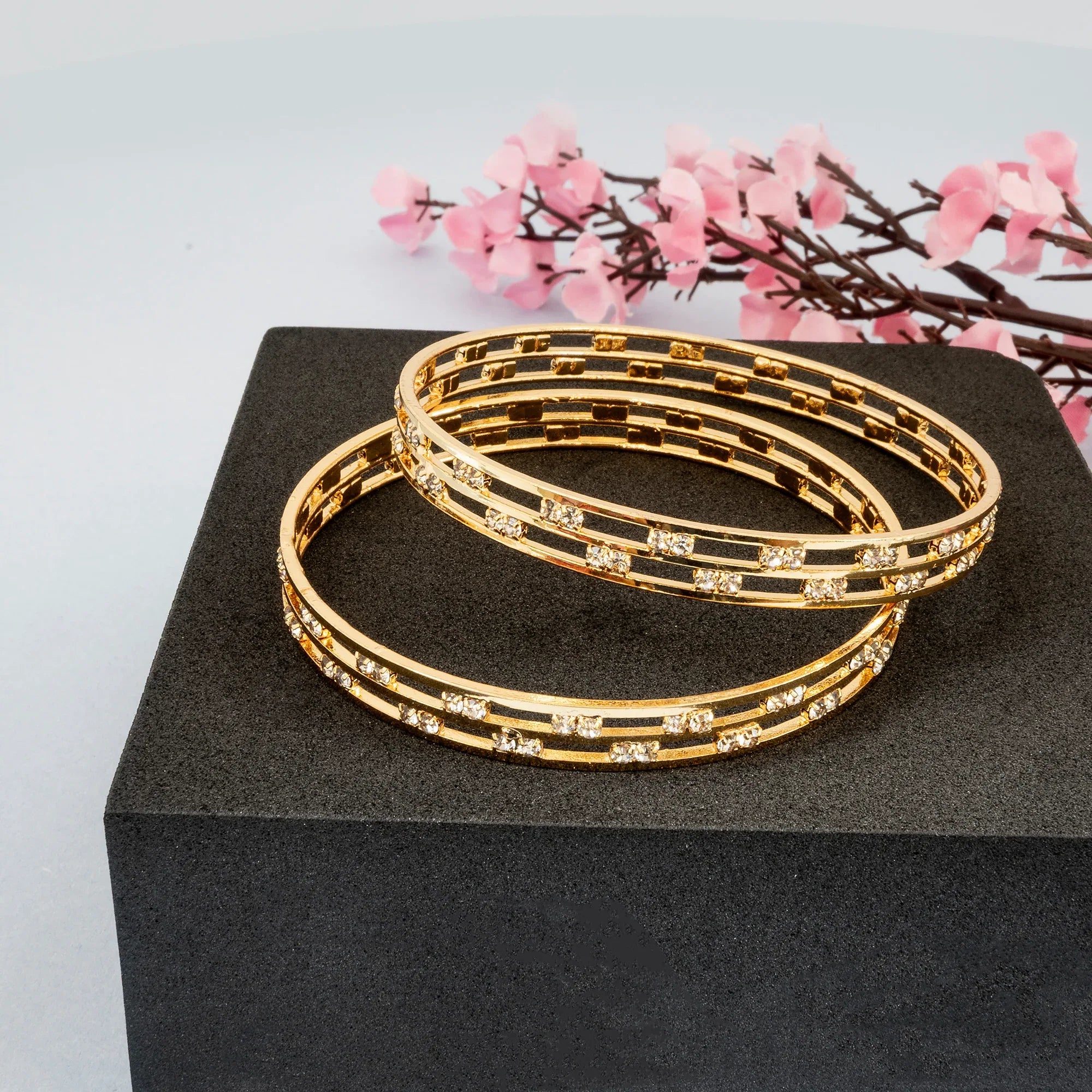 Gold bangles with crystal accents on a black stand with pink flowers in the background