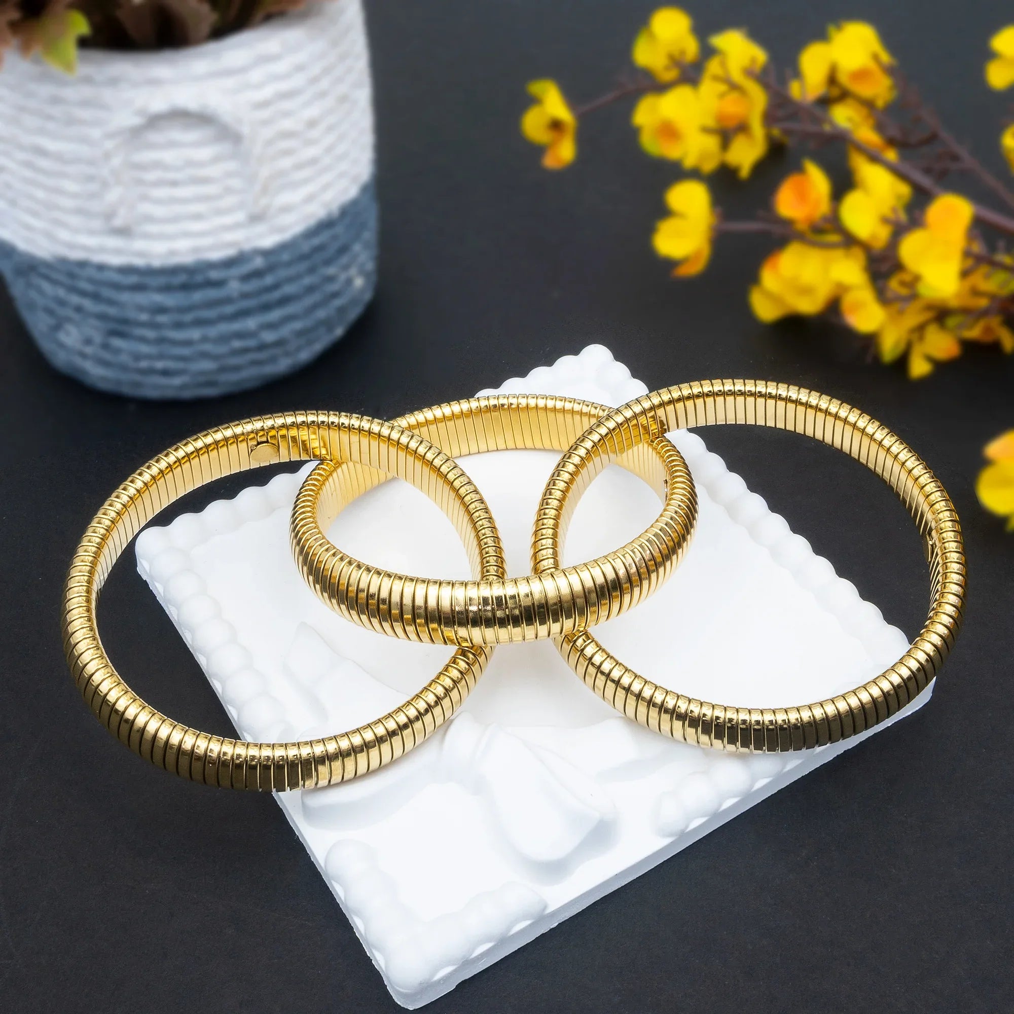 Three gold bangles on a white cushion with yellow flowers in the background
