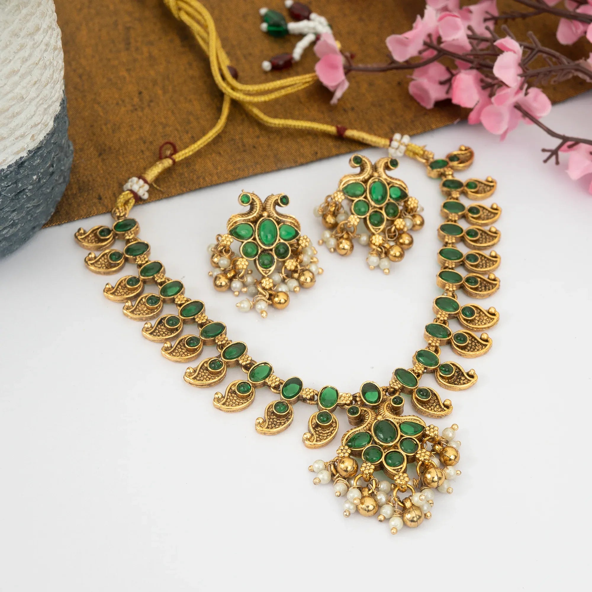 Gold and green jewelry set with earrings and necklace on a white surface with pink flowers.