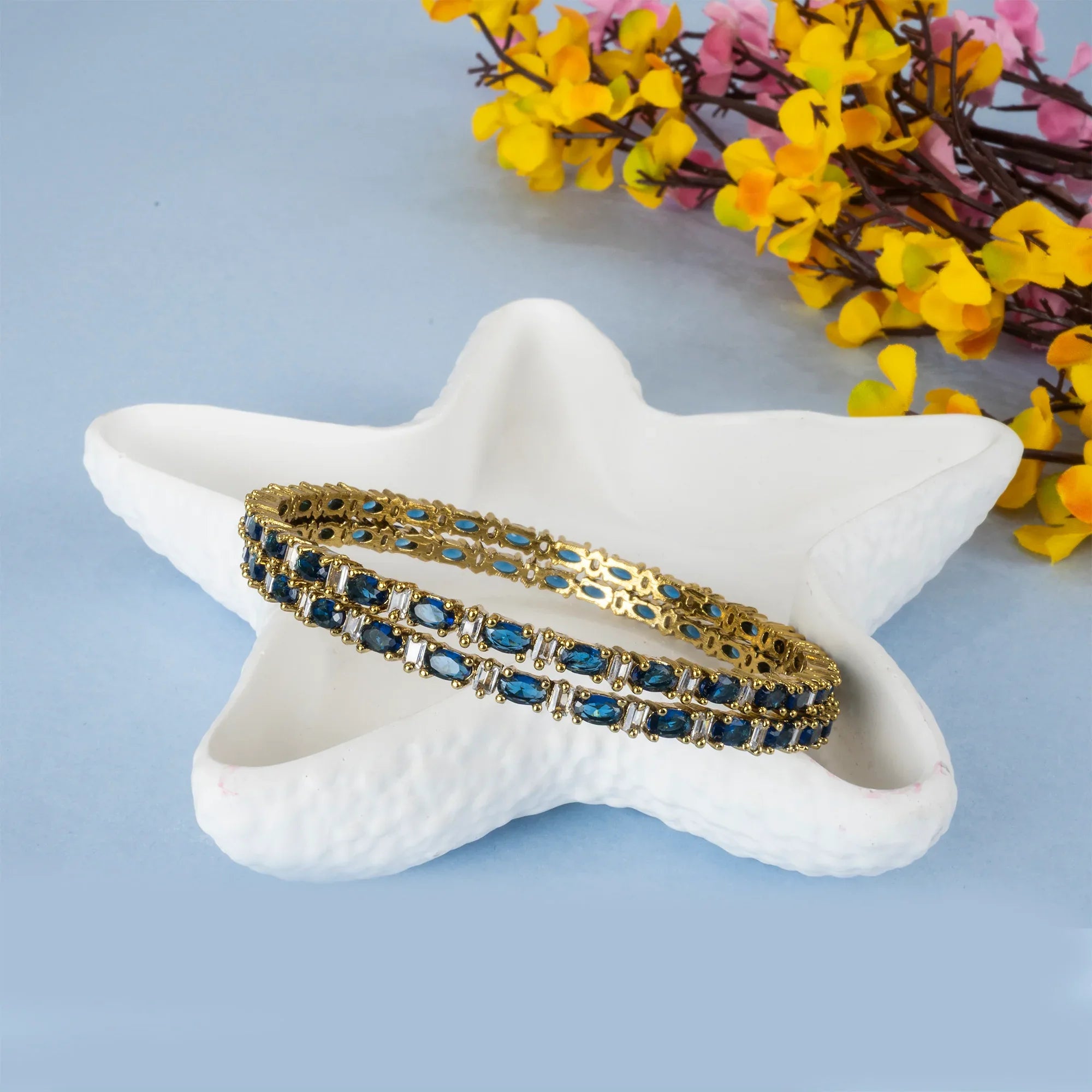Gold and blue bracelet on a star-shaped white dish with flowers in the background