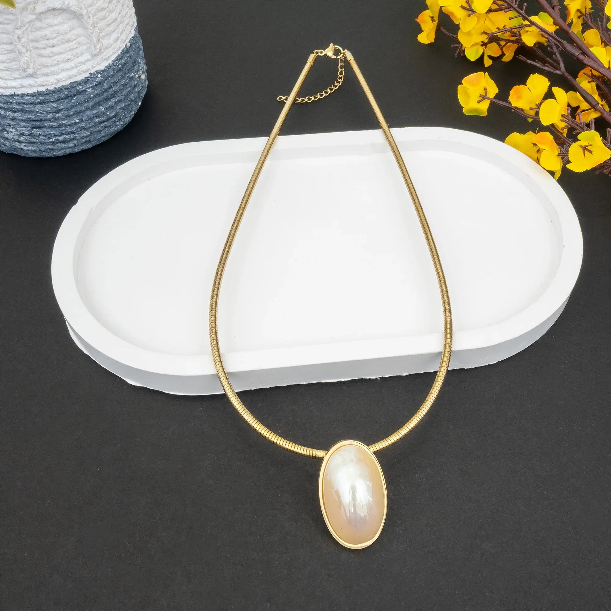 Gold necklace with a large pearl pendant on a dark surface with yellow flowers and a woven basket.