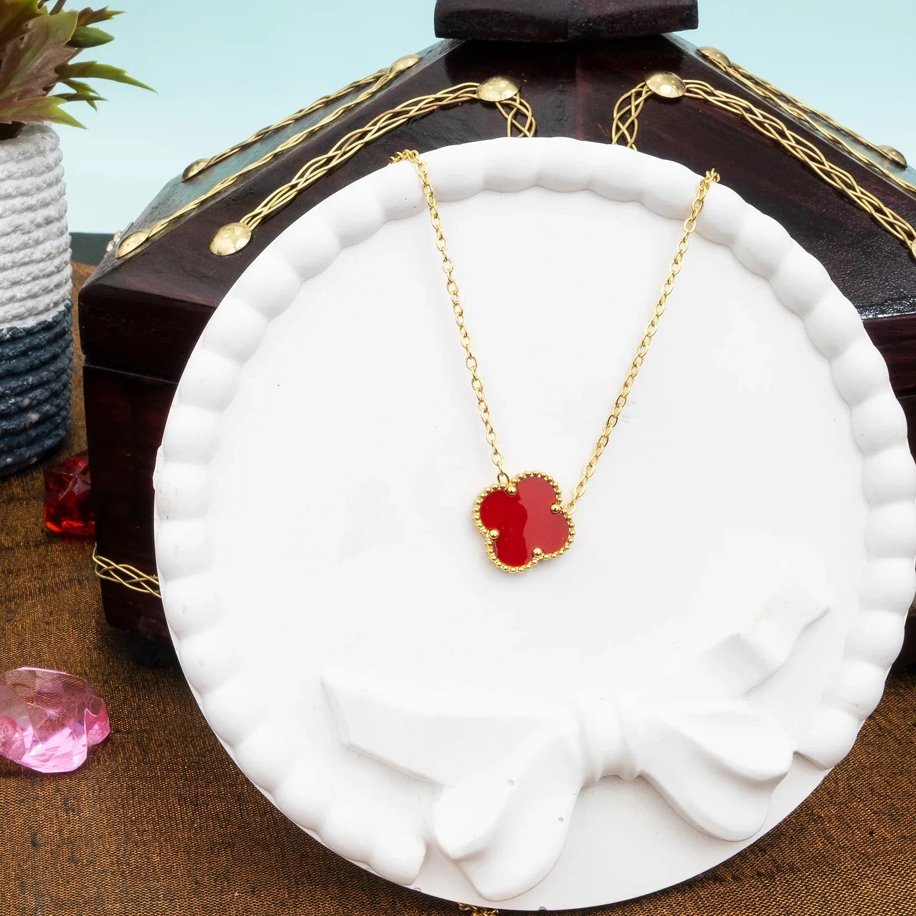 Gold necklace with a red pendant on a white stand, with a wooden box and pink gemstone in the background.