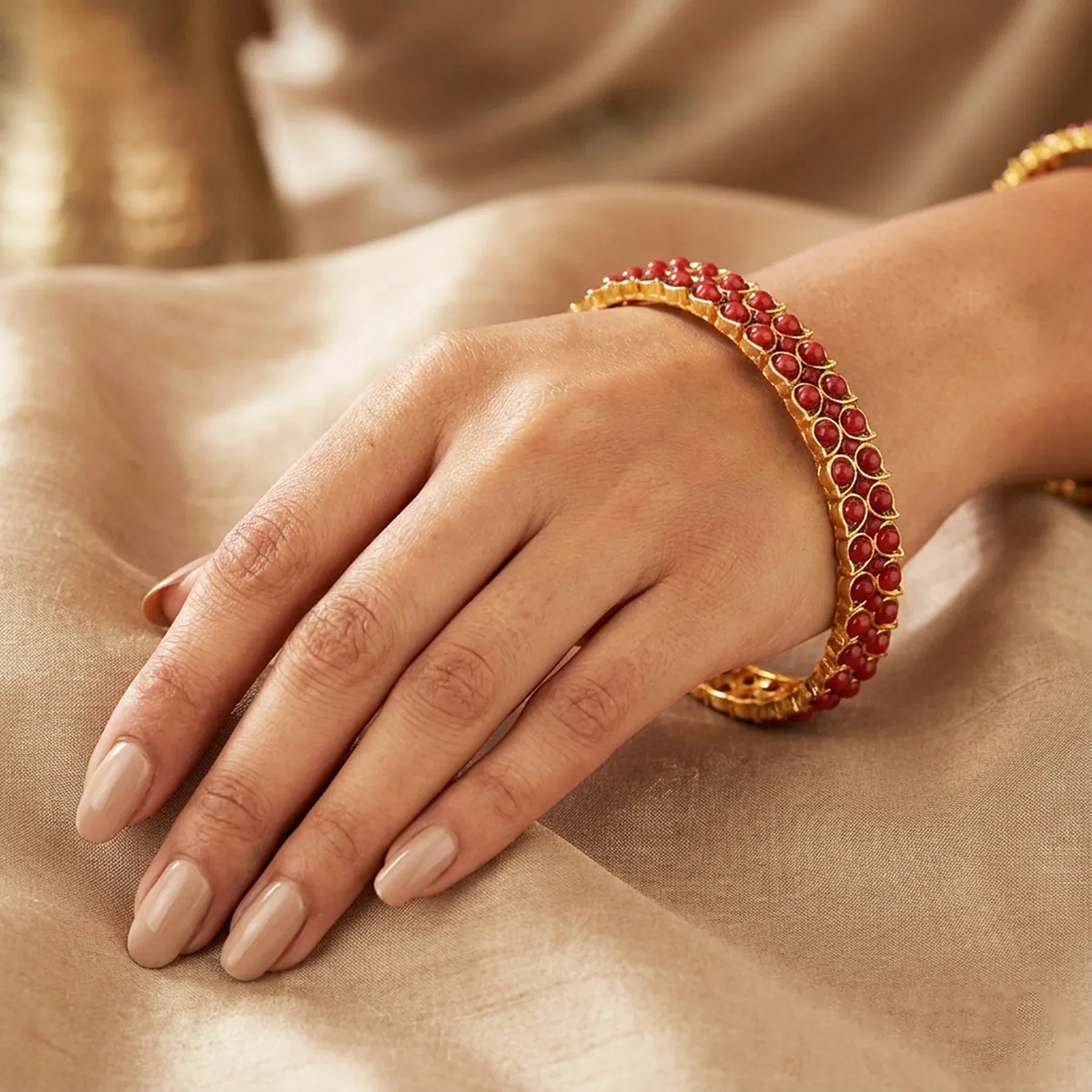 Hand wearing a gold bracelet with red stones on a beige fabric background