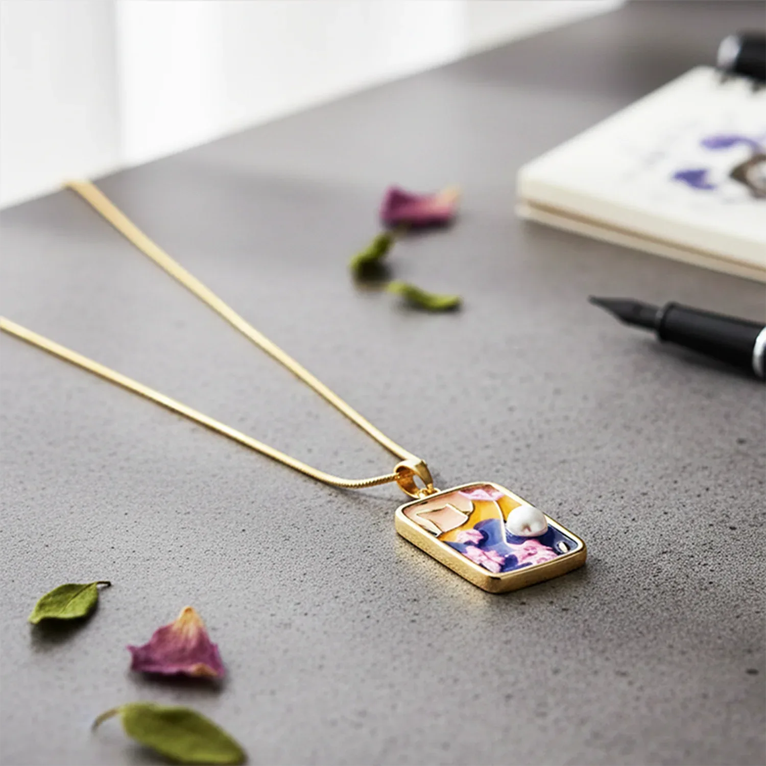 Gold necklace with a rectangular pendant on a gray surface with dried flowers and a pen.