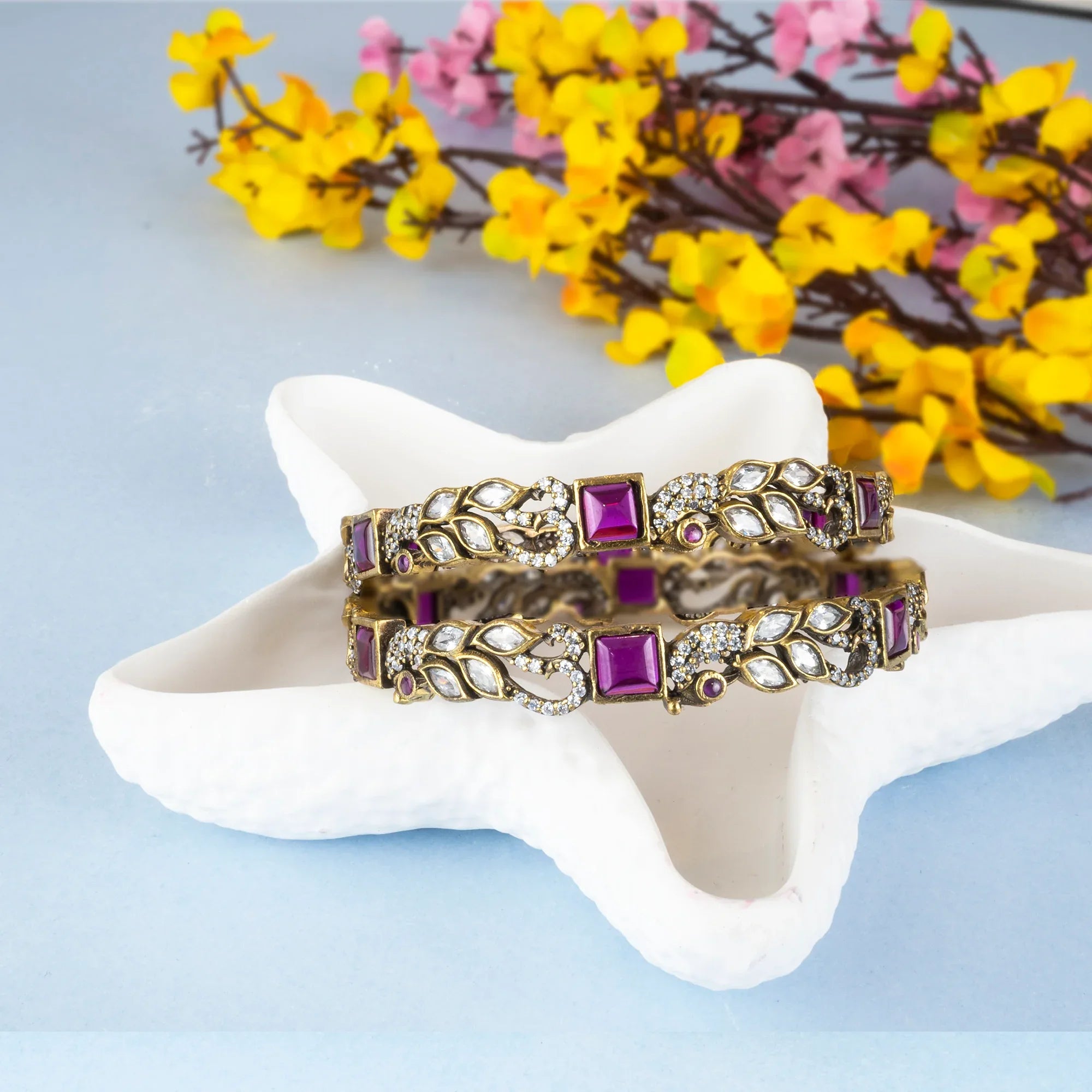 Two decorative bracelets with purple stones on a white star-shaped stand with flowers in the background.