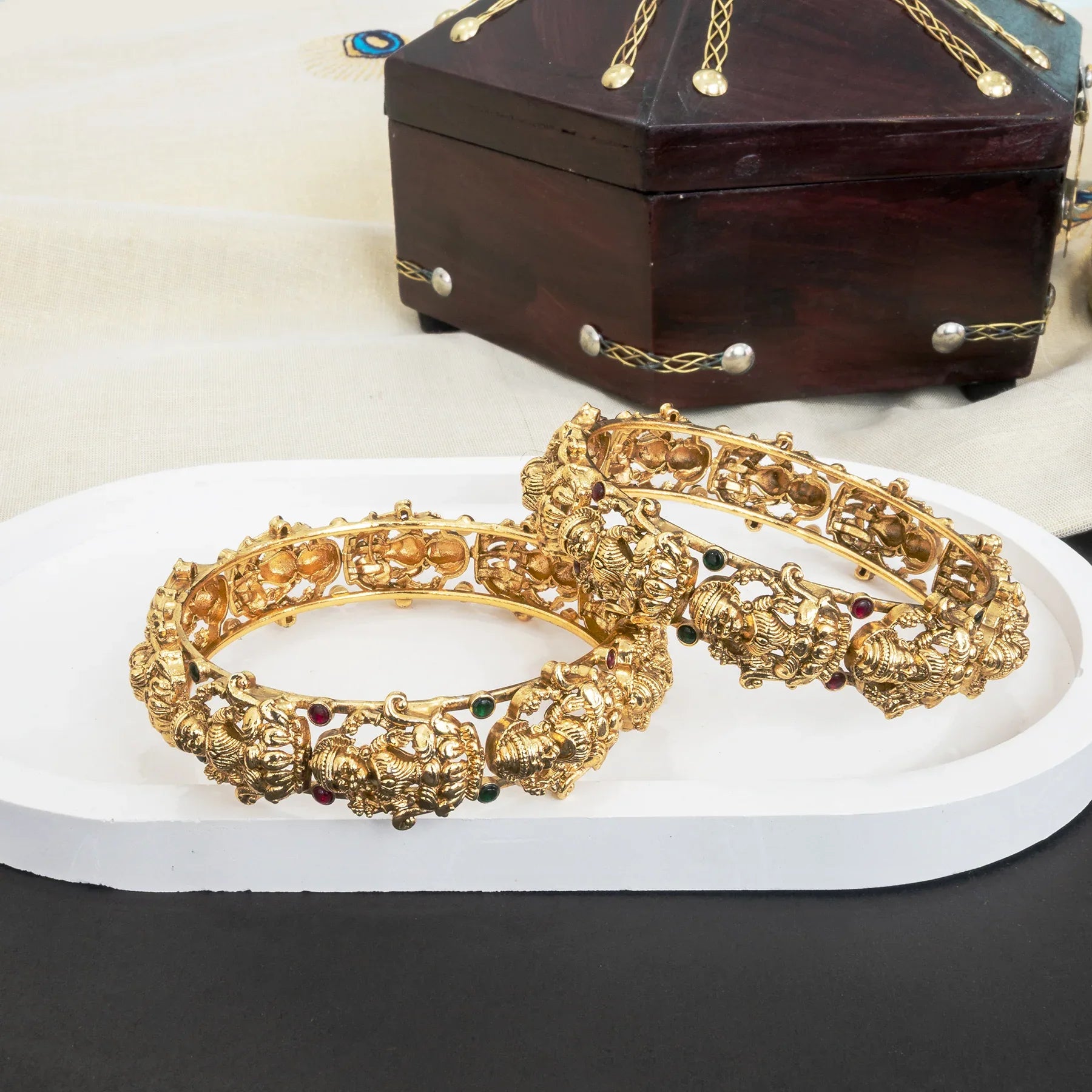 Gold bangles with intricate designs on a white platform, with a wooden jewelry box in the background.