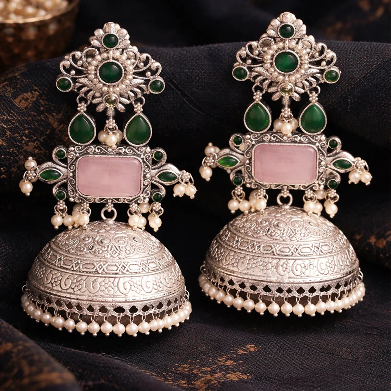 Pair of ornate silver earrings with green gemstones and pink stones on a dark fabric background