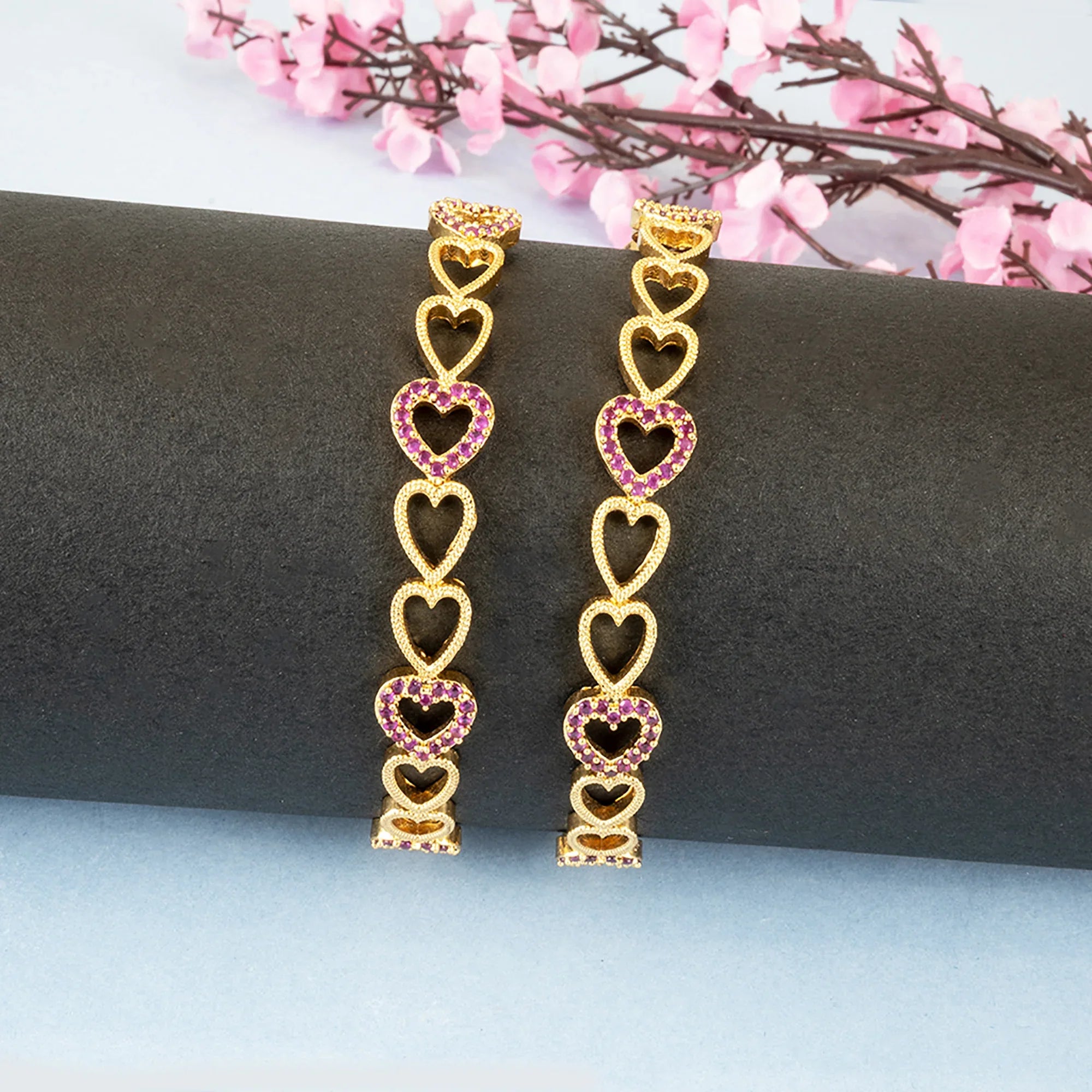 Gold heart-shaped earrings with pink gemstones on a dark surface with pink flowers in the background.
