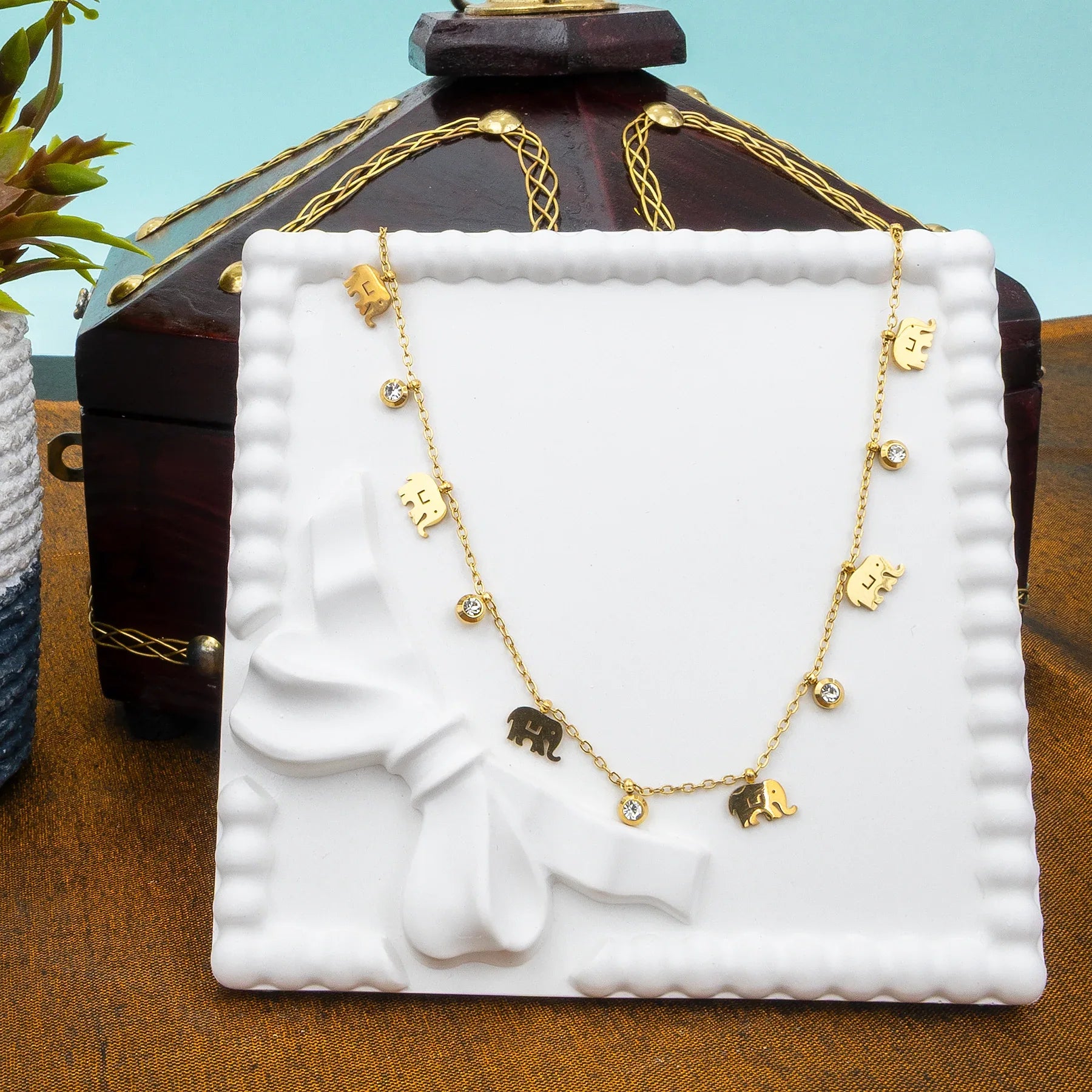 Gold necklace with elephant charms on a white stand with a wooden box and plant in the background