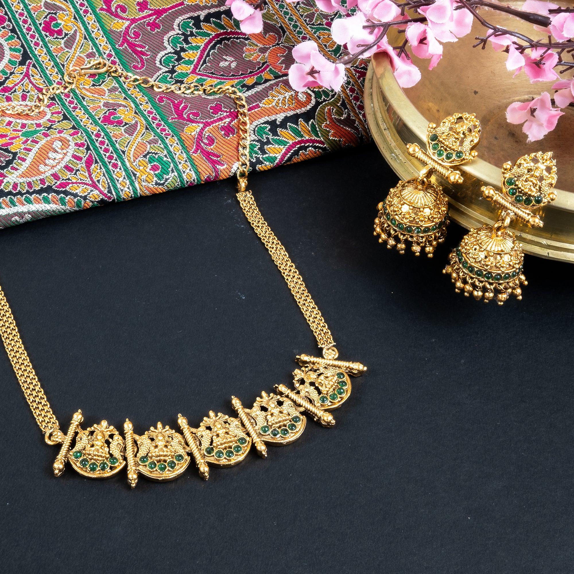 Gold necklace and earrings on a dark surface with a colorful fabric and pink flowers in the background.