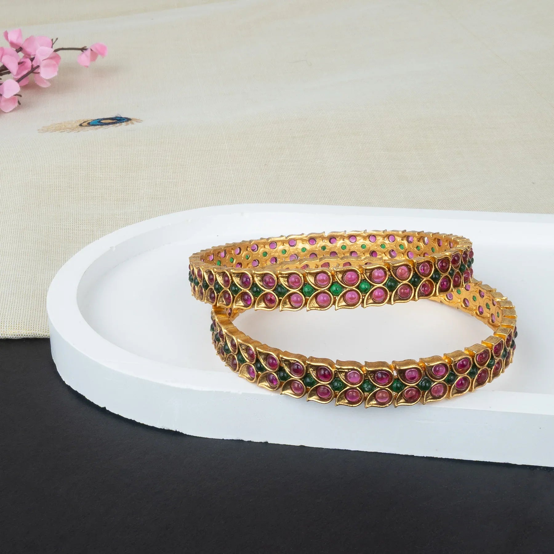 Pair of gold bangles with pink and green stones on a white platform.