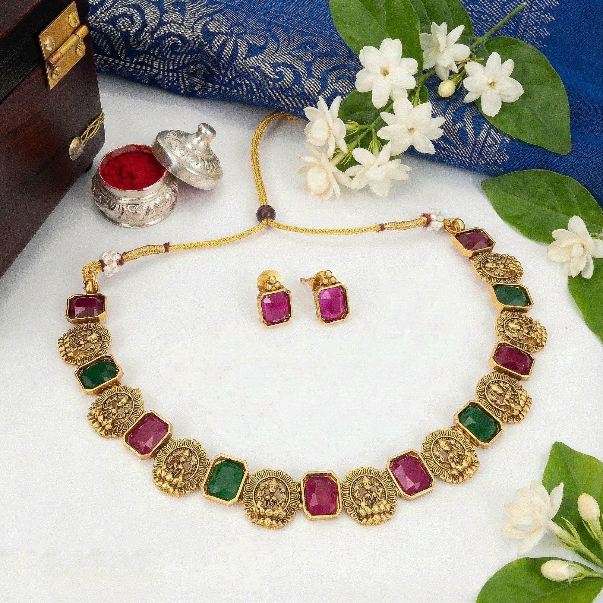 Gold necklace with red and green gemstones on a white surface with flowers and jewelry box.