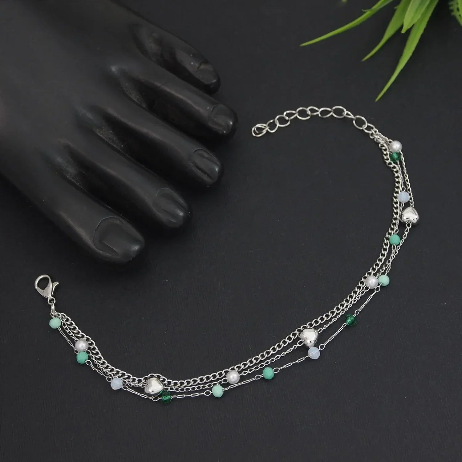 Multistrand bracelet with pearls and green beads on a dark surface with a hand and plant in the background.