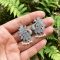 Peacock-shaped earrings with pink and green stones held in a hand against a natural background.