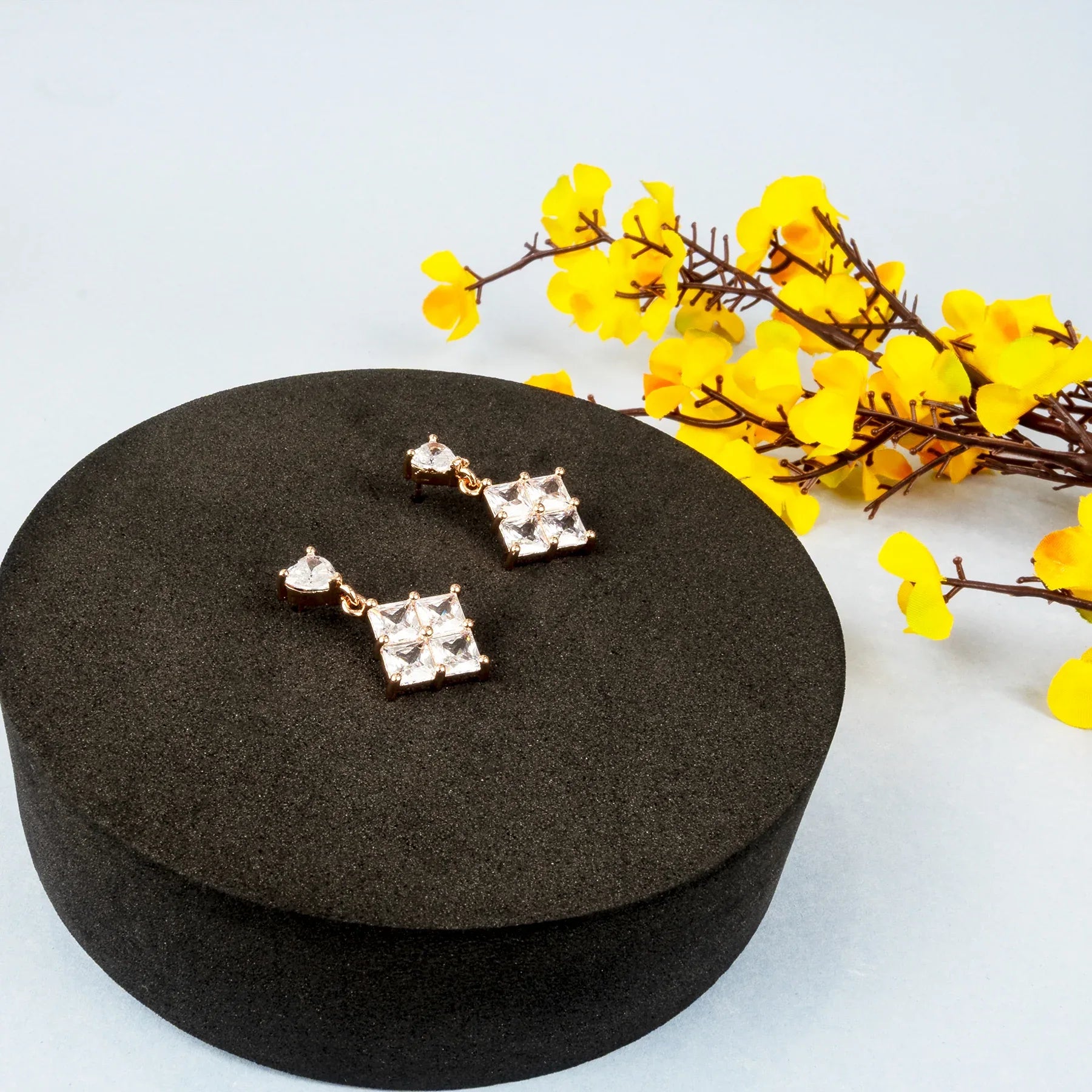 Pair of square-shaped earrings on a black round stand with yellow flowers in the background