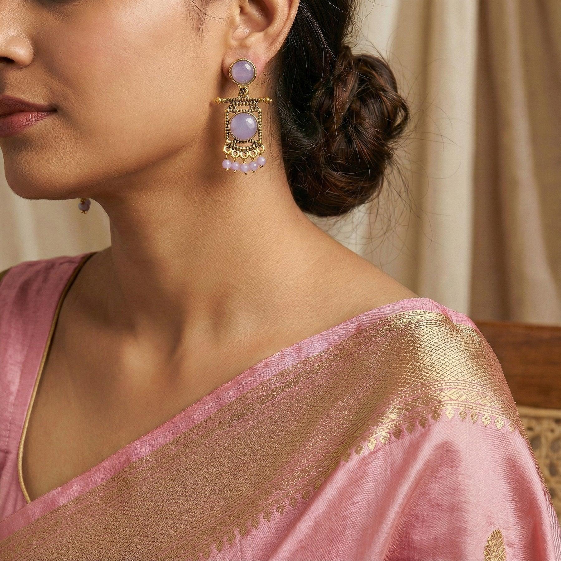 Woman wearing a pink saree with gold border and earrings with purple stones.