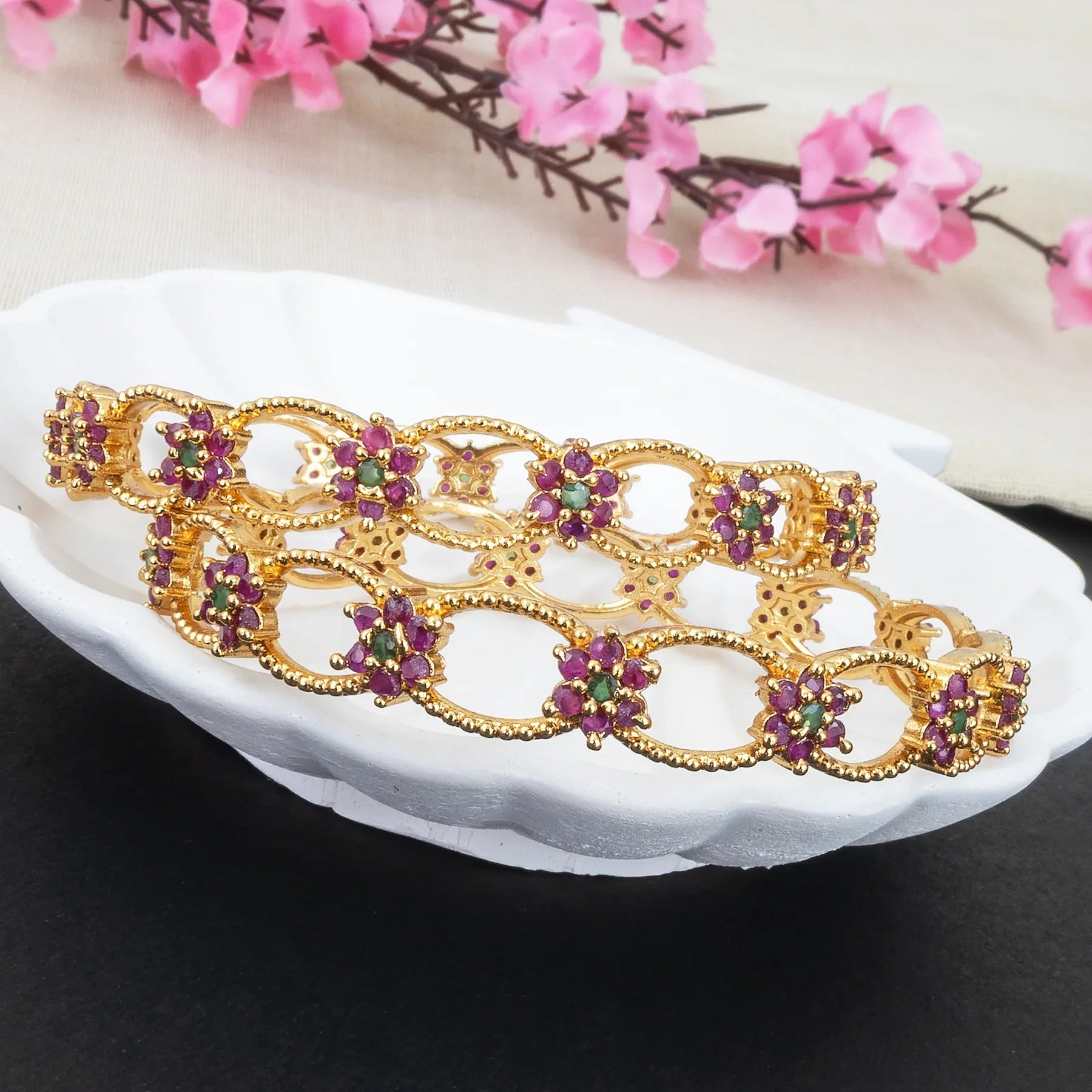 Gold bracelet with purple and green gemstones on a white dish with pink flowers in the background