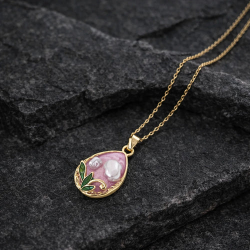 Gold necklace with a pendant featuring a pink stone and green leaves on a dark stone surface