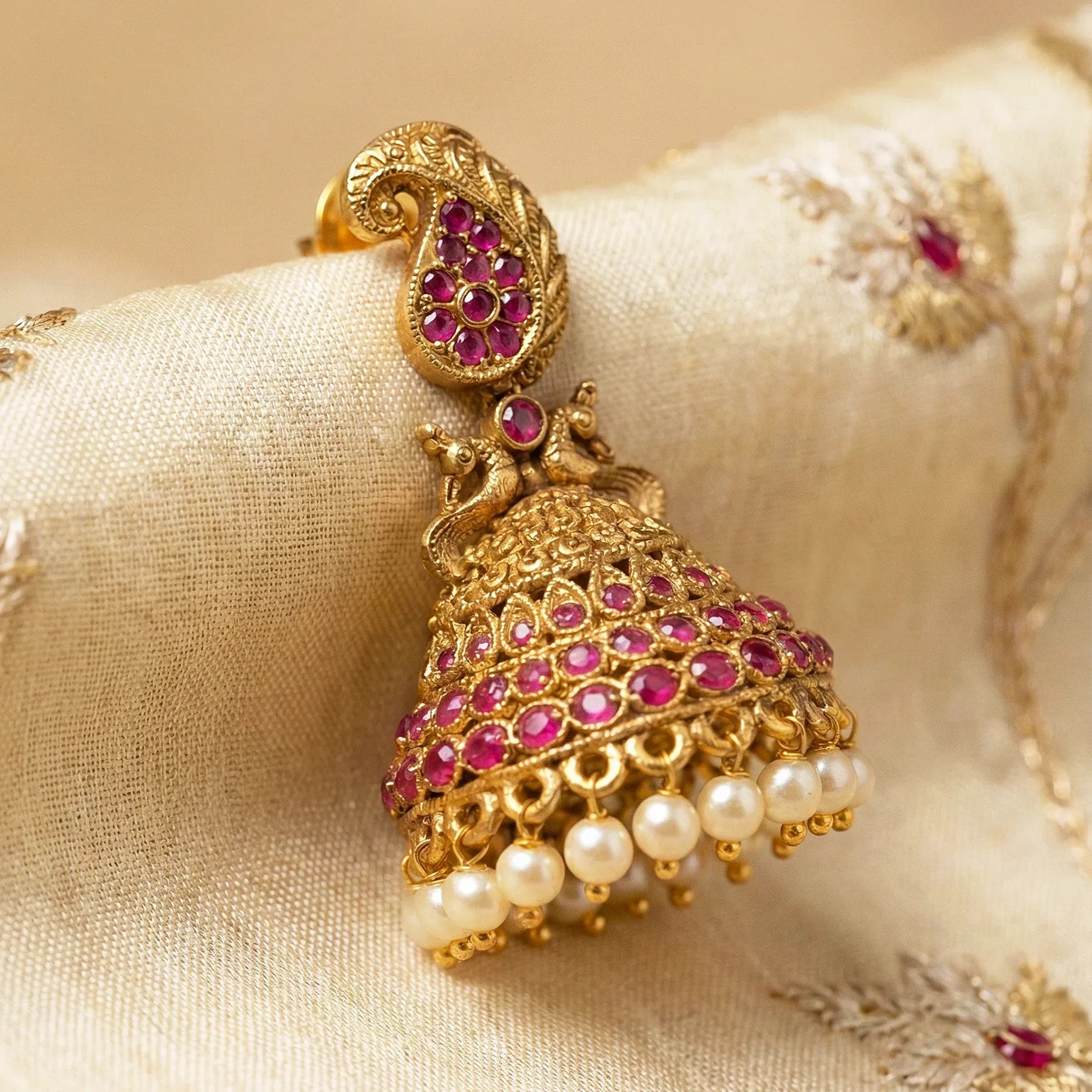 Ornate gold earring with pink gemstones and pearls on a beige fabric background