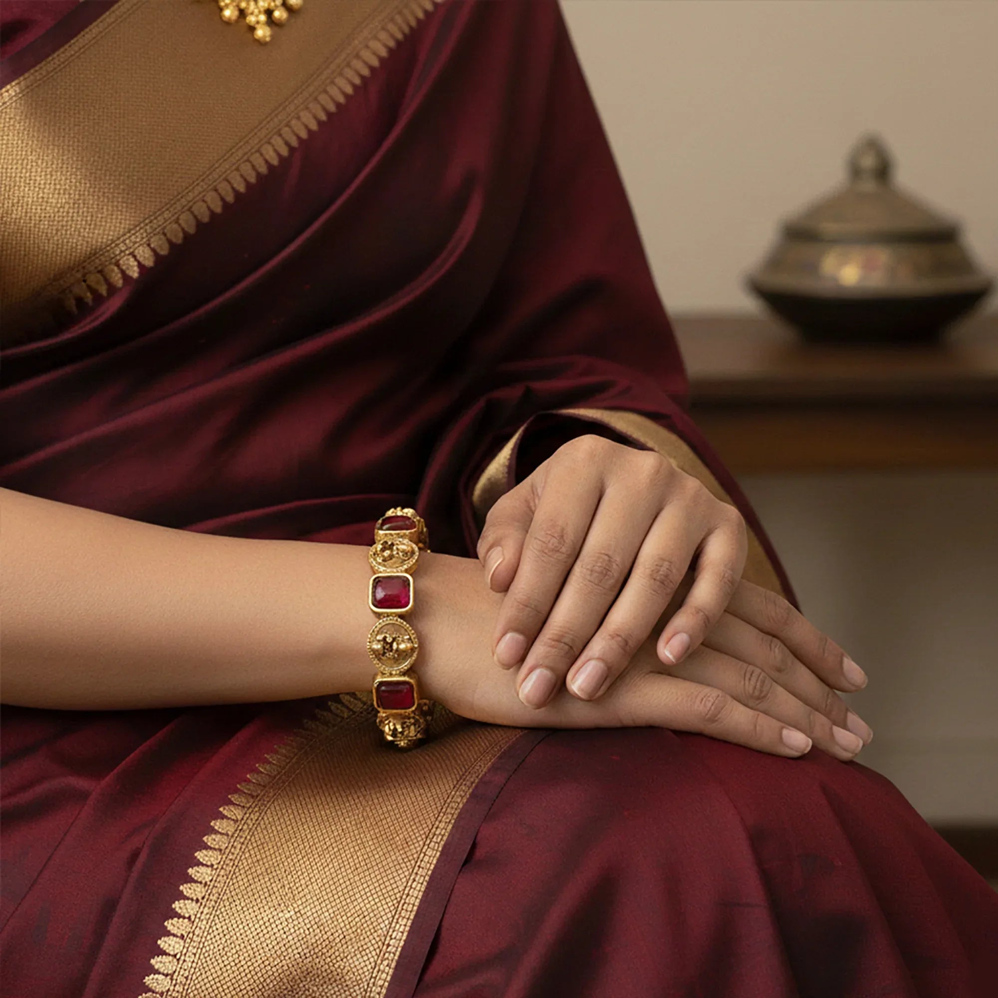 Person wearing a gold bracelet with red stones on a maroon saree.