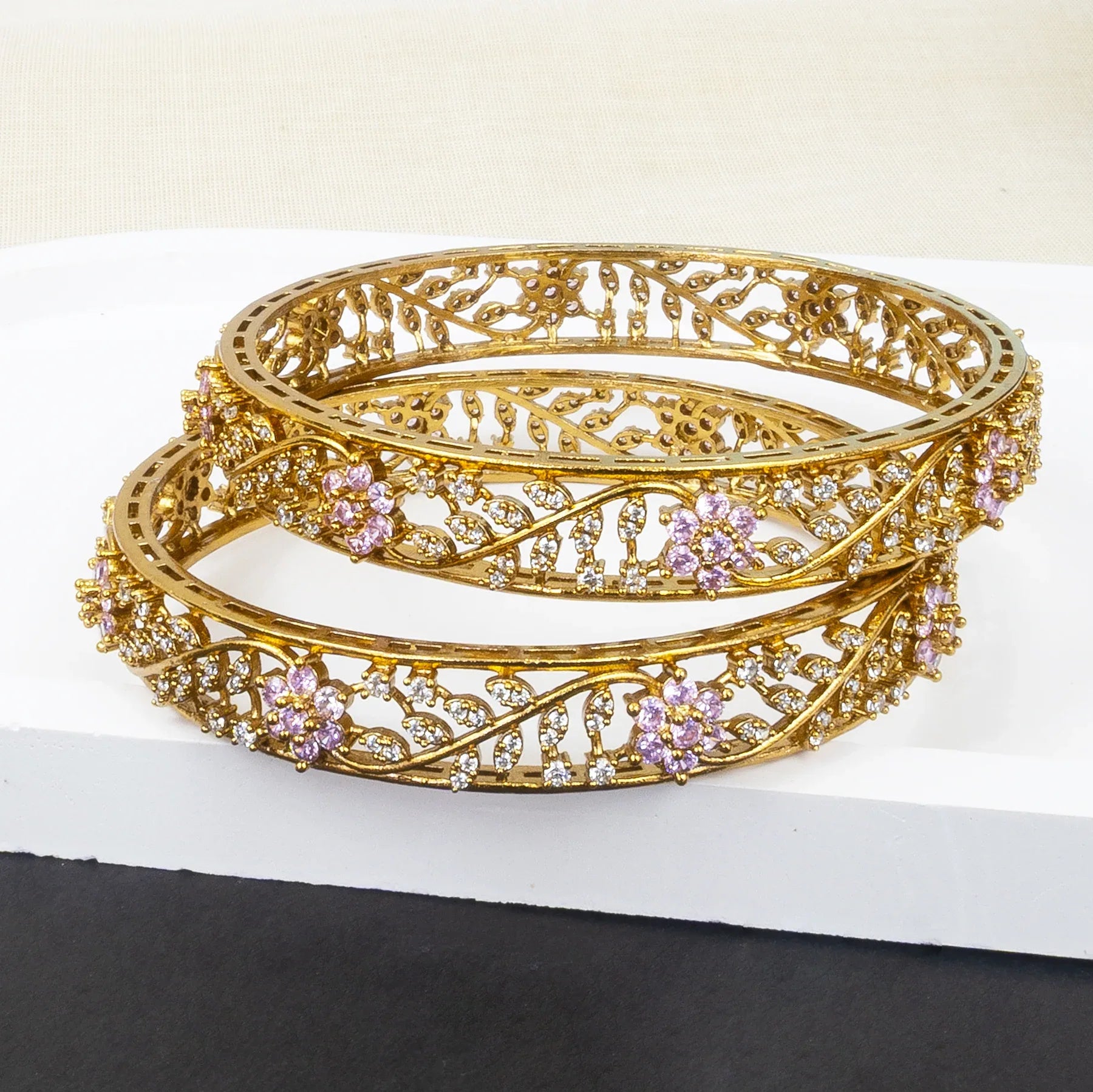 Gold bangles with intricate designs and pink stones on a white surface