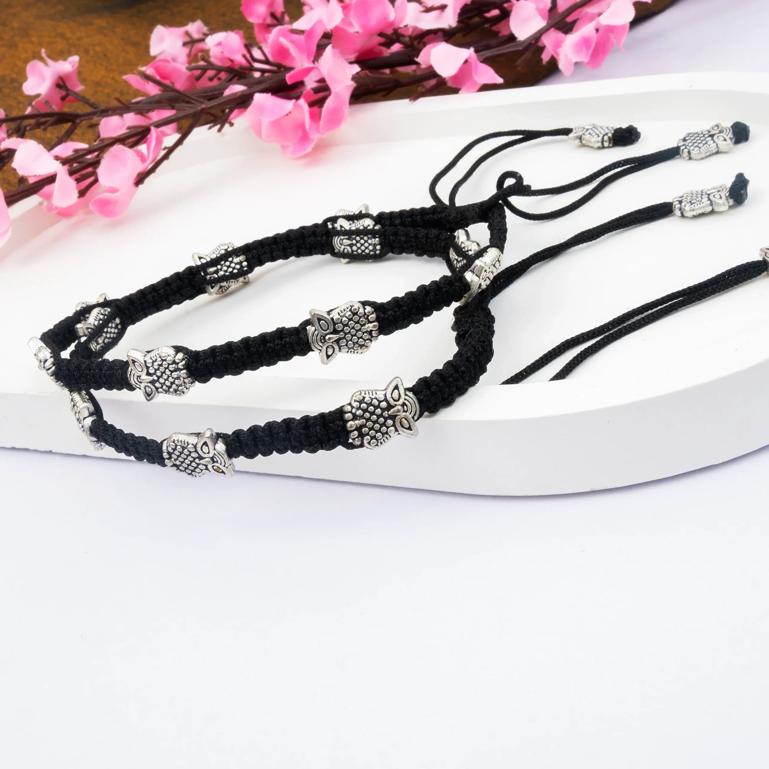 Black braided bracelets with silver owl charms on a white surface with pink flowers in the background